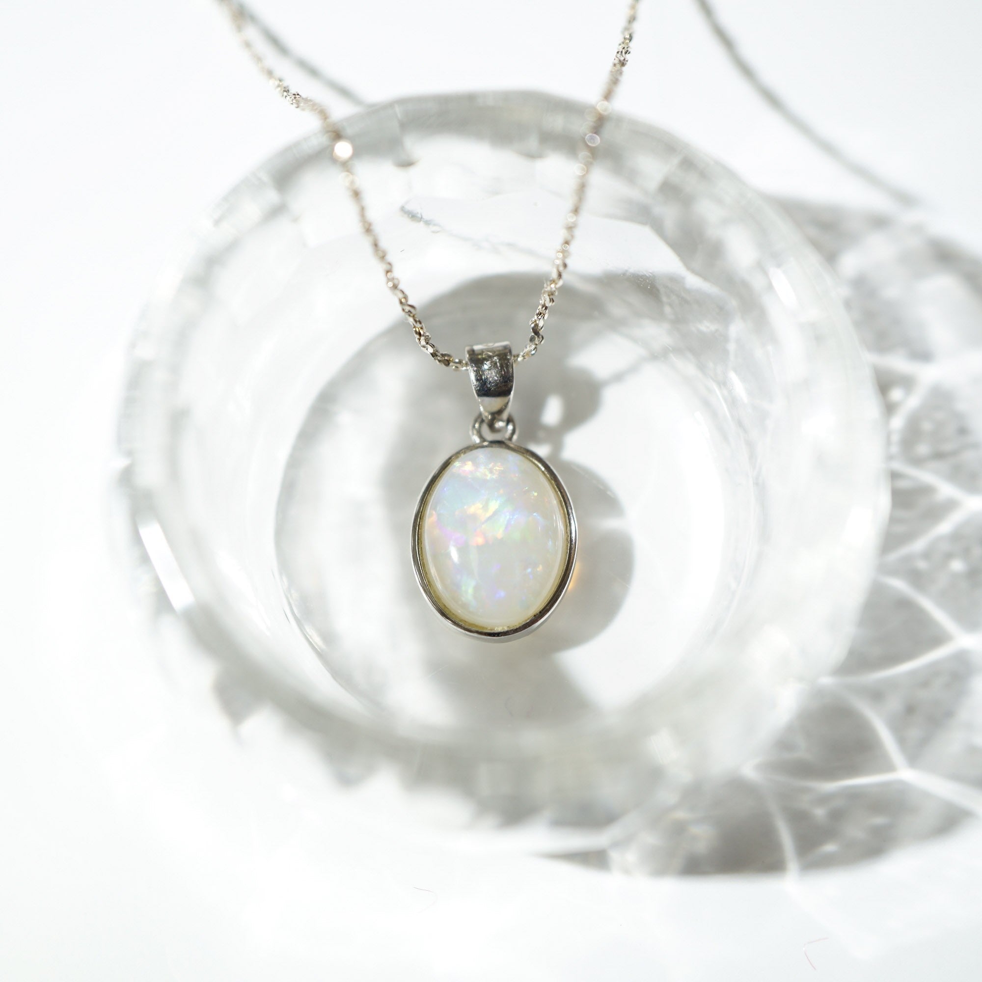 A silver pendant necklace with an oval iridescent opal stone, resting on a clear glass dish against a bright white background.