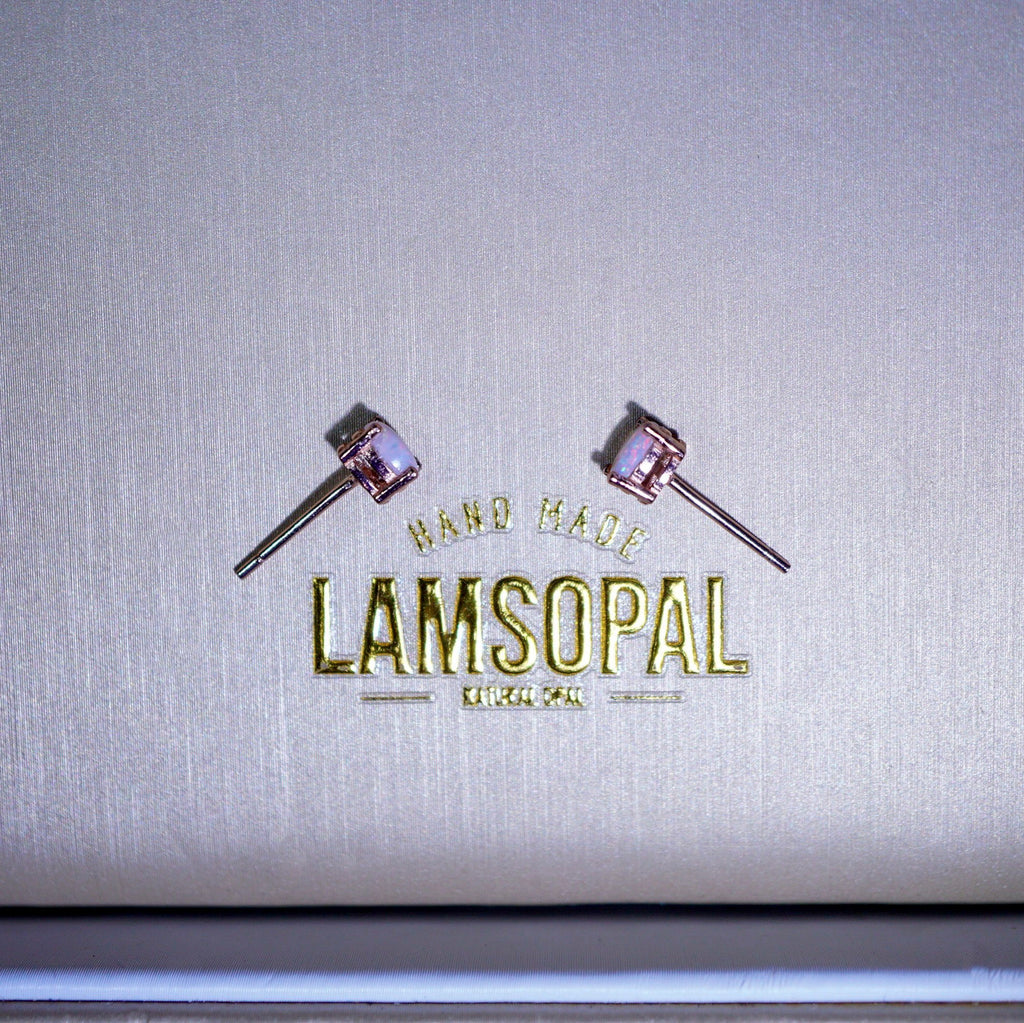 A pair of small stud earrings with pinkish-purple cabochon stones and silver-toned posts, displayed on a textured metallic silver surface above a gold-foiled Lamsopal logo.