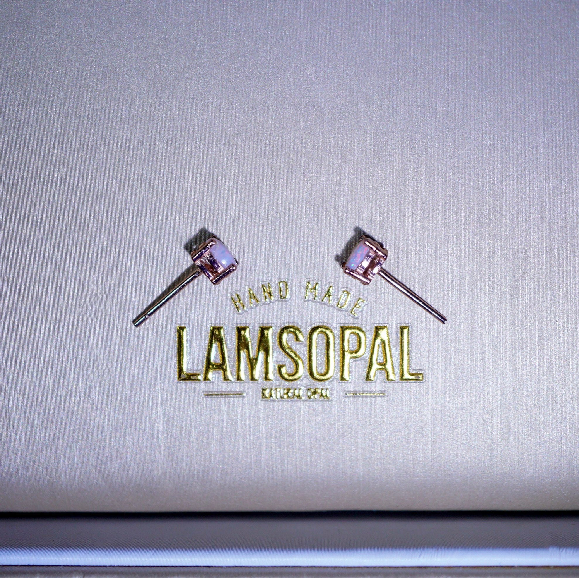 A pair of small stud earrings with pinkish-purple cabochon stones and silver-toned posts, displayed on a textured metallic silver surface above a gold-foiled Lamsopal logo.