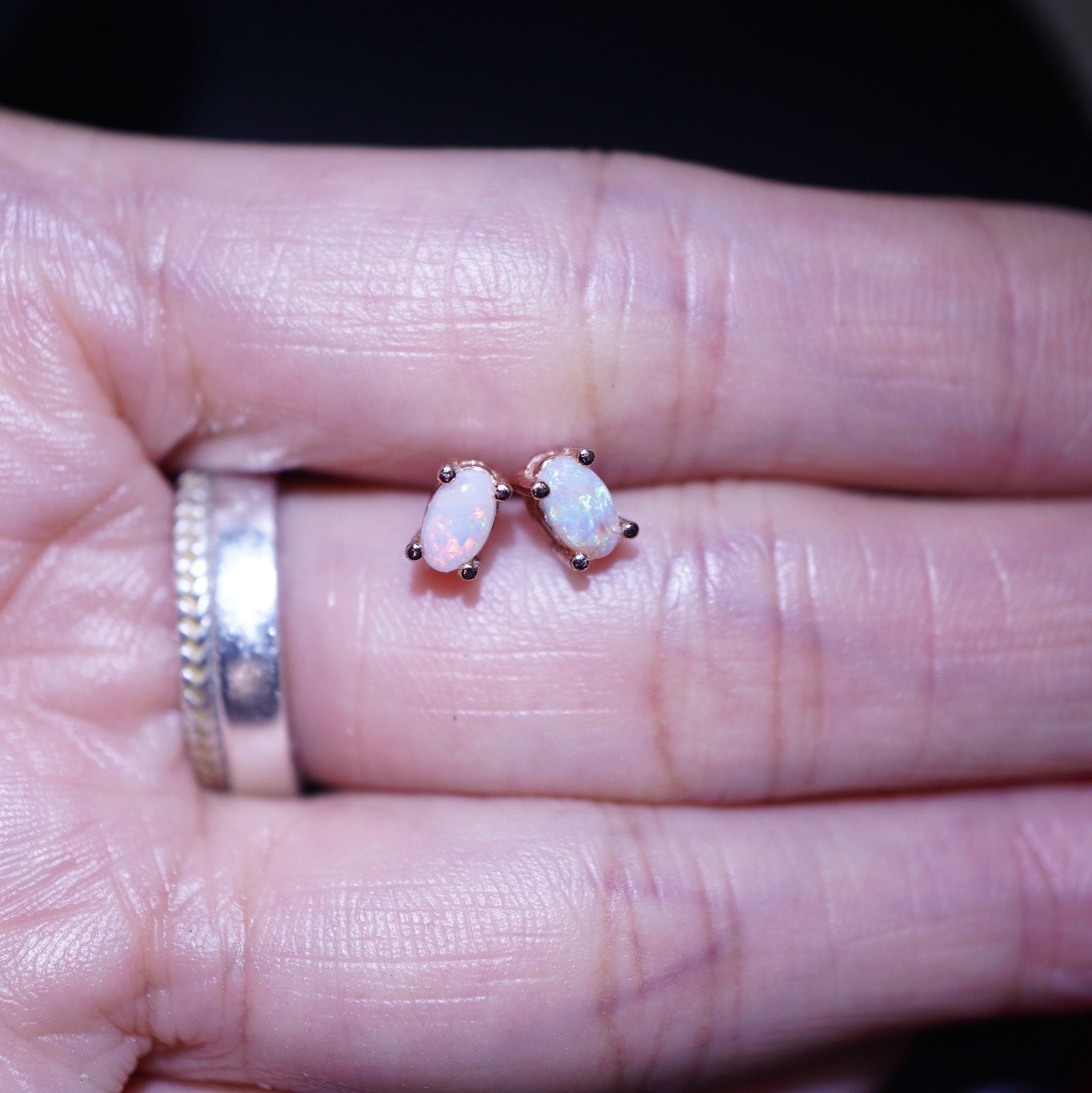 A close-up of a person's hand wearing a pair of small oval stud earrings with iridescent white opal-like centers in rose-gold-toned settings, alongside a silver ring.