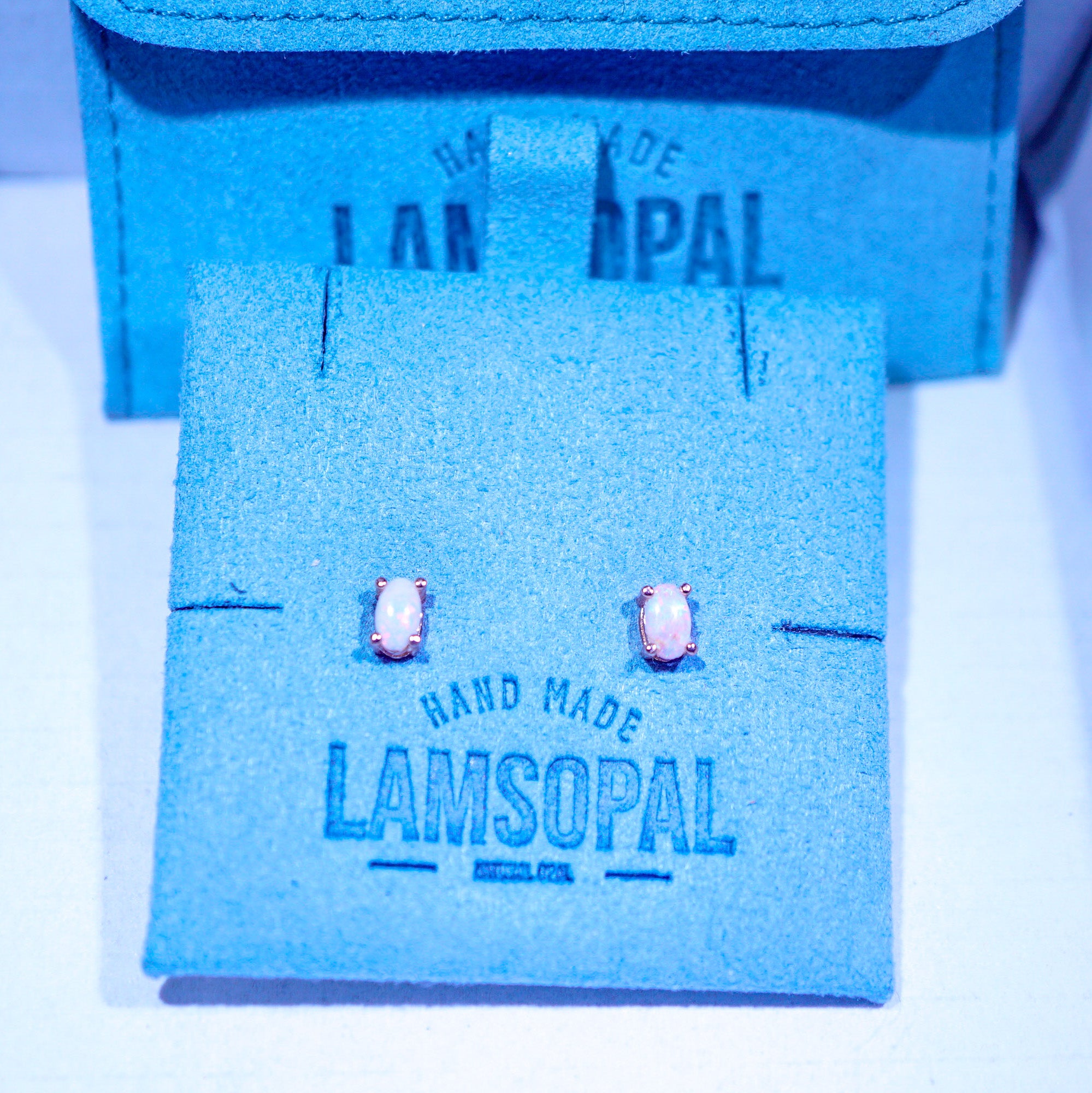 A pair of small oval stud earrings with pale pink stones in silver-toned settings, displayed on a blue suede jewelry card branded Lamsopal, with the matching box behind it.