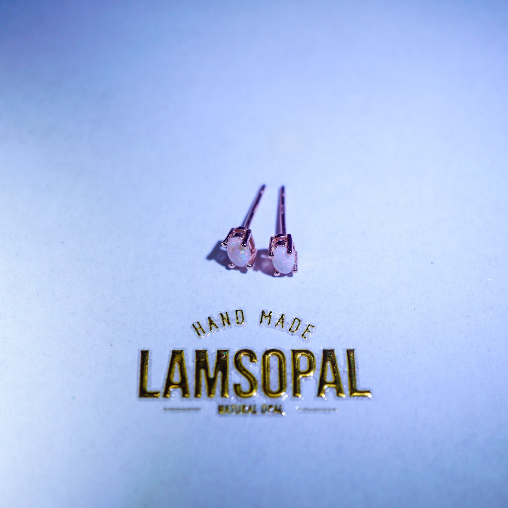 A pair of small stud earrings with pinkish-purple stones and silver-toned posts, placed above gold foil text reading 'HAND MADE LAMSOPAL' on a light blue background.