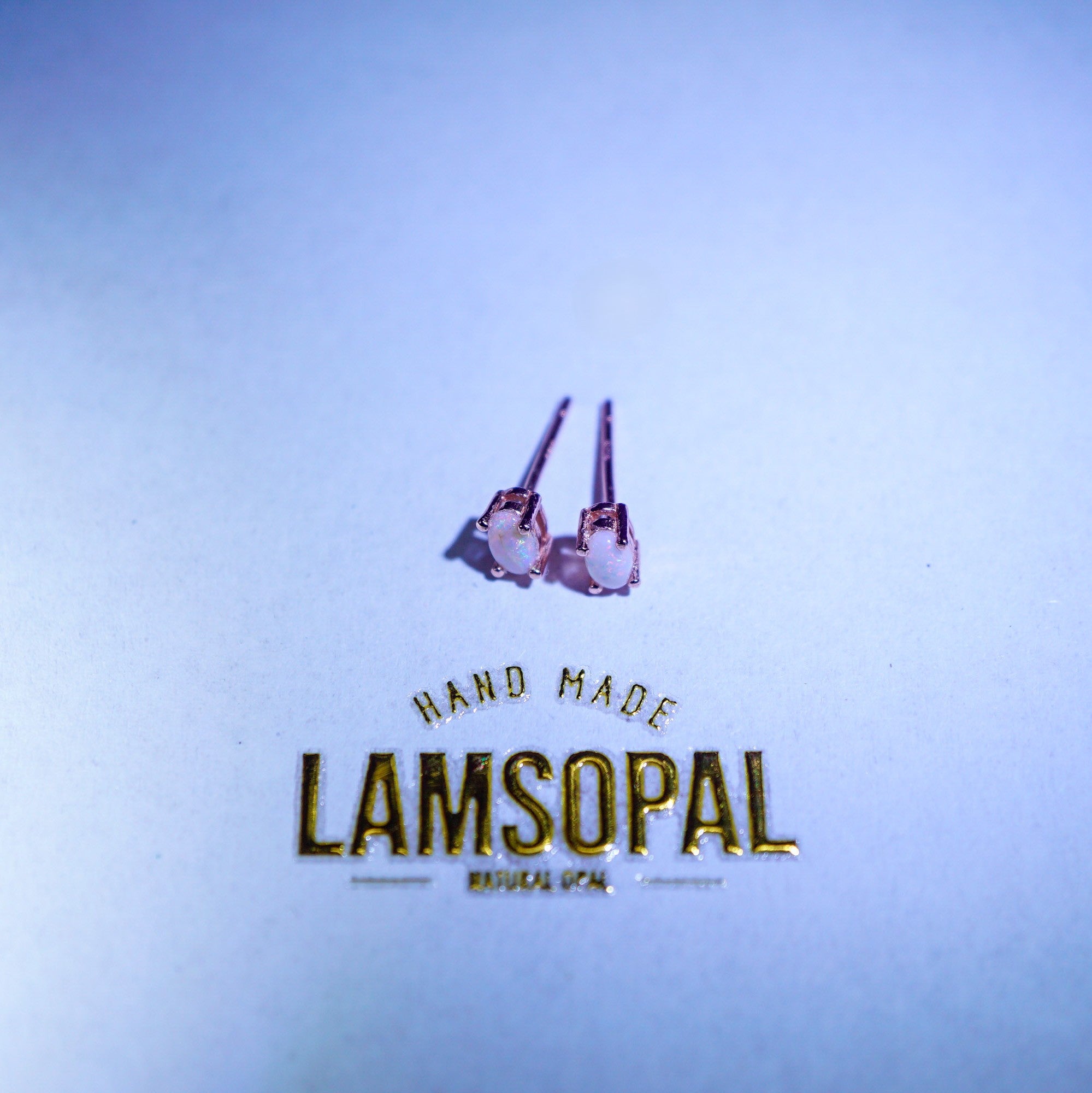 A pair of small stud earrings with pinkish-purple stones and silver-toned posts, placed above gold foil text reading 'HAND MADE LAMSOPAL' on a light blue background.