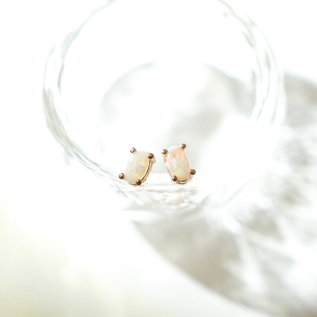 A pair of small oval stud earrings with iridescent opal-like stones in a gold-tone setting, resting inside a clear glass dish on a bright white surface.