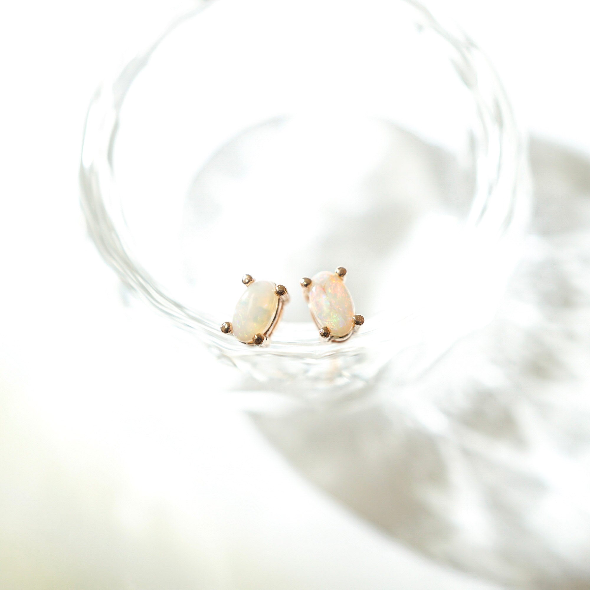 A pair of small oval stud earrings with iridescent opal-like stones in a gold-tone setting, resting inside a clear glass dish on a bright white surface.