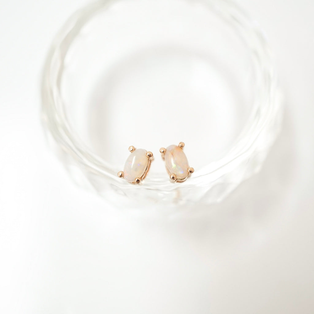 A pair of rose gold stud earrings with oval iridescent opal gemstones, resting on the rim of a clear glass tumbler against a bright white background.