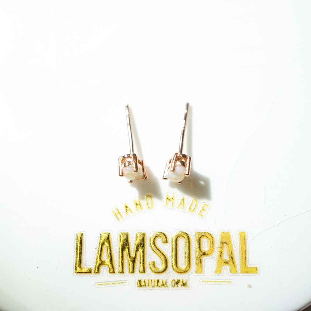 A pair of small stud earrings with rose gold-toned metal settings and oval natural opal stones, displayed on a white surface above gold foil text reading 'HAND MADE LAMSOPAL NATURAL OPAL'.