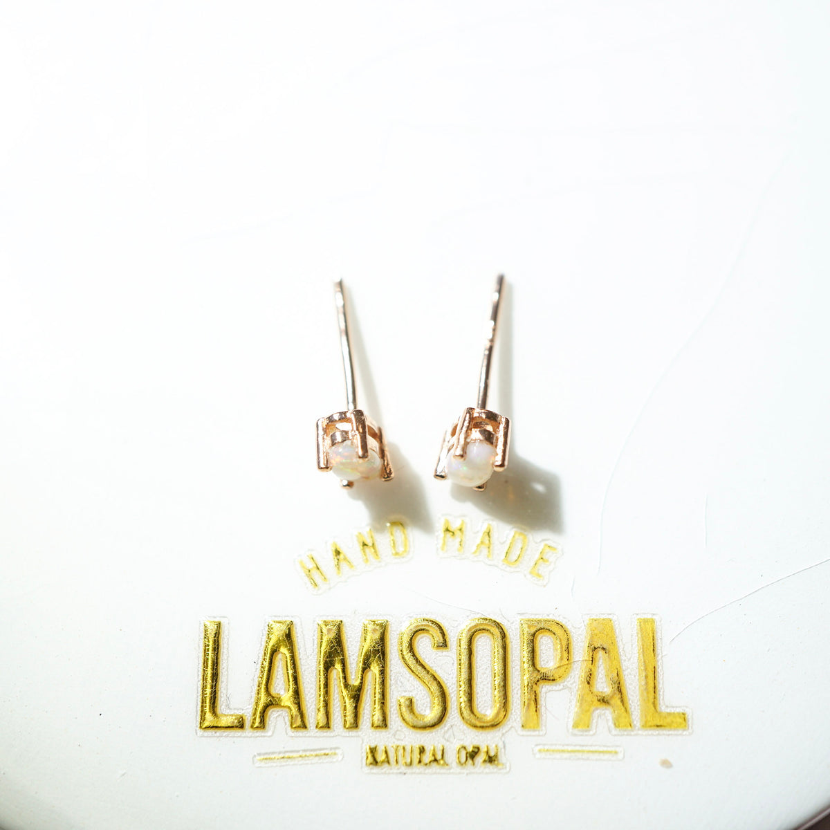 A pair of small stud earrings with rose gold-toned metal settings and oval natural opal stones, displayed on a white surface above gold foil text reading 'HAND MADE LAMSOPAL NATURAL OPAL'.
