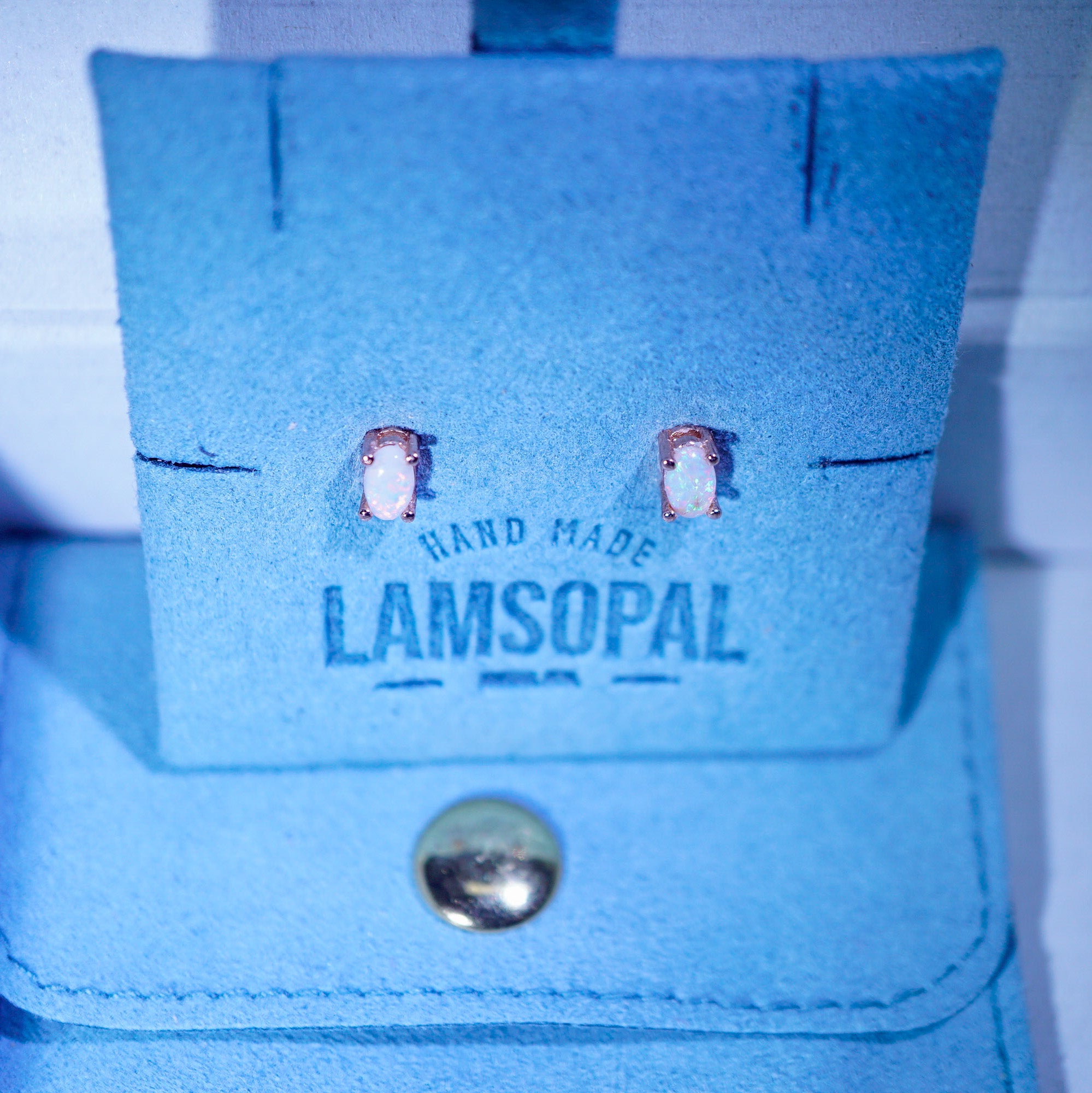 A pair of small stud earrings with pale pink stones set in rose gold, displayed on a blue suede stand with the brand name Lamsopal printed on it.