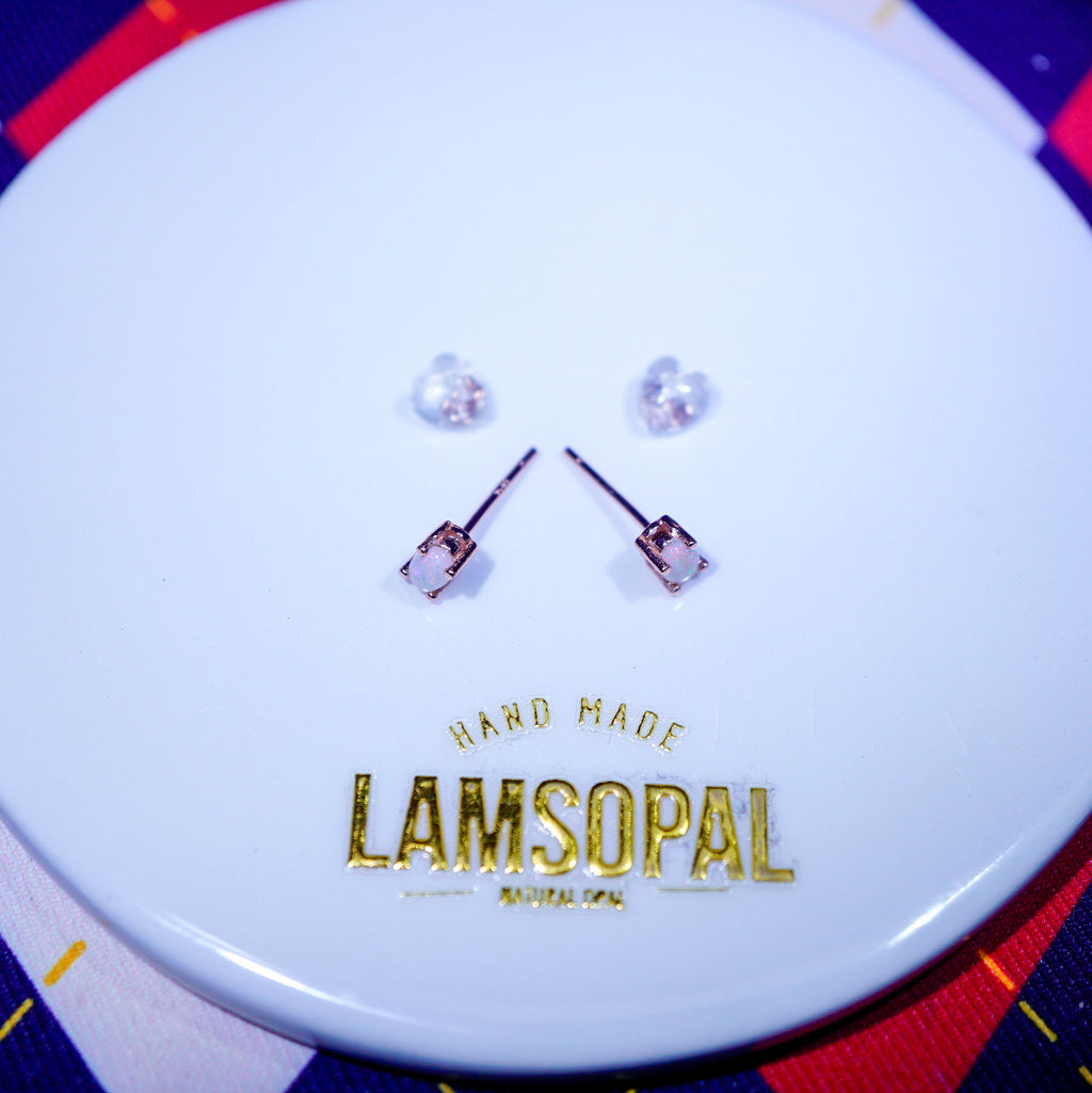 A pair of small stud earrings with clear faceted stones and rose-gold-toned settings, displayed on a white circular card printed with the brand name LAMSOPAL.