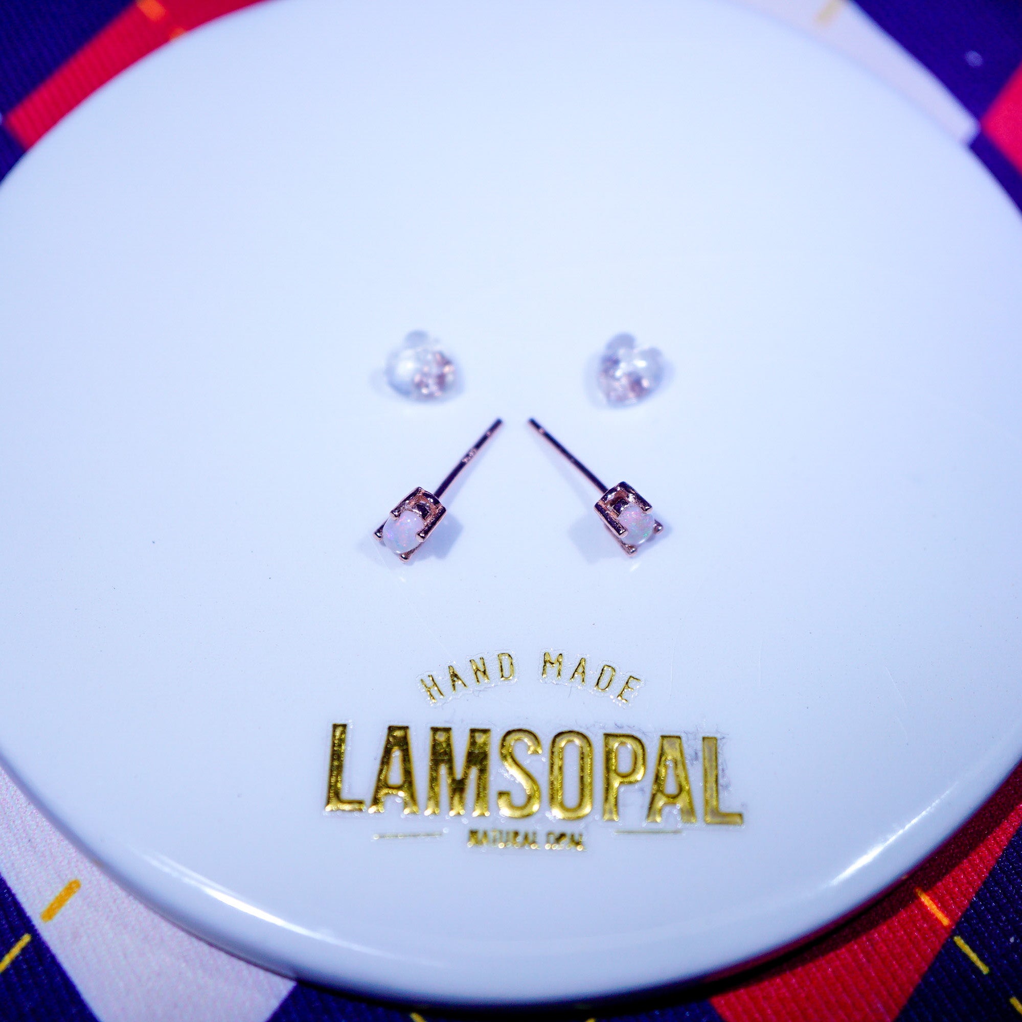 A pair of small stud earrings with clear faceted stones and rose-gold-toned settings, displayed on a white circular card printed with the brand name LAMSOPAL.