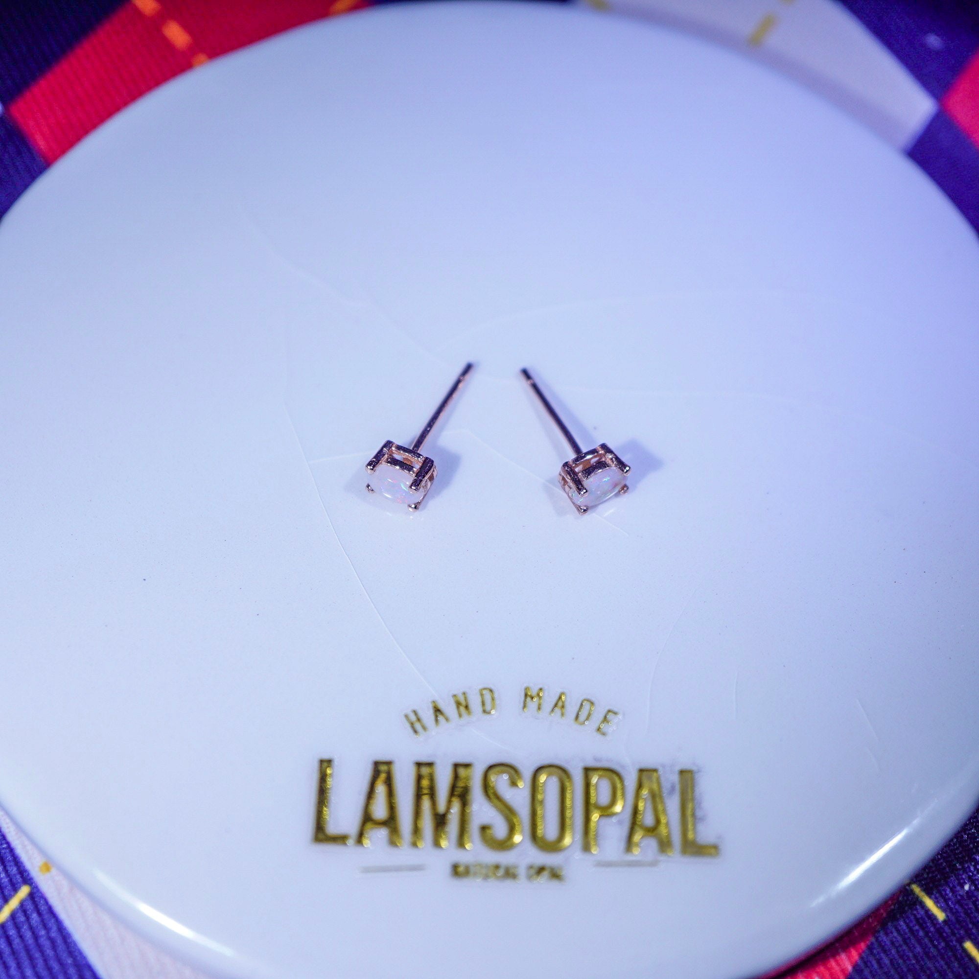 A pair of small stud earrings with clear square-cut stones in silver-toned settings, displayed on a white circular branded card that reads 'HAND MADE LAMSOPAL'.