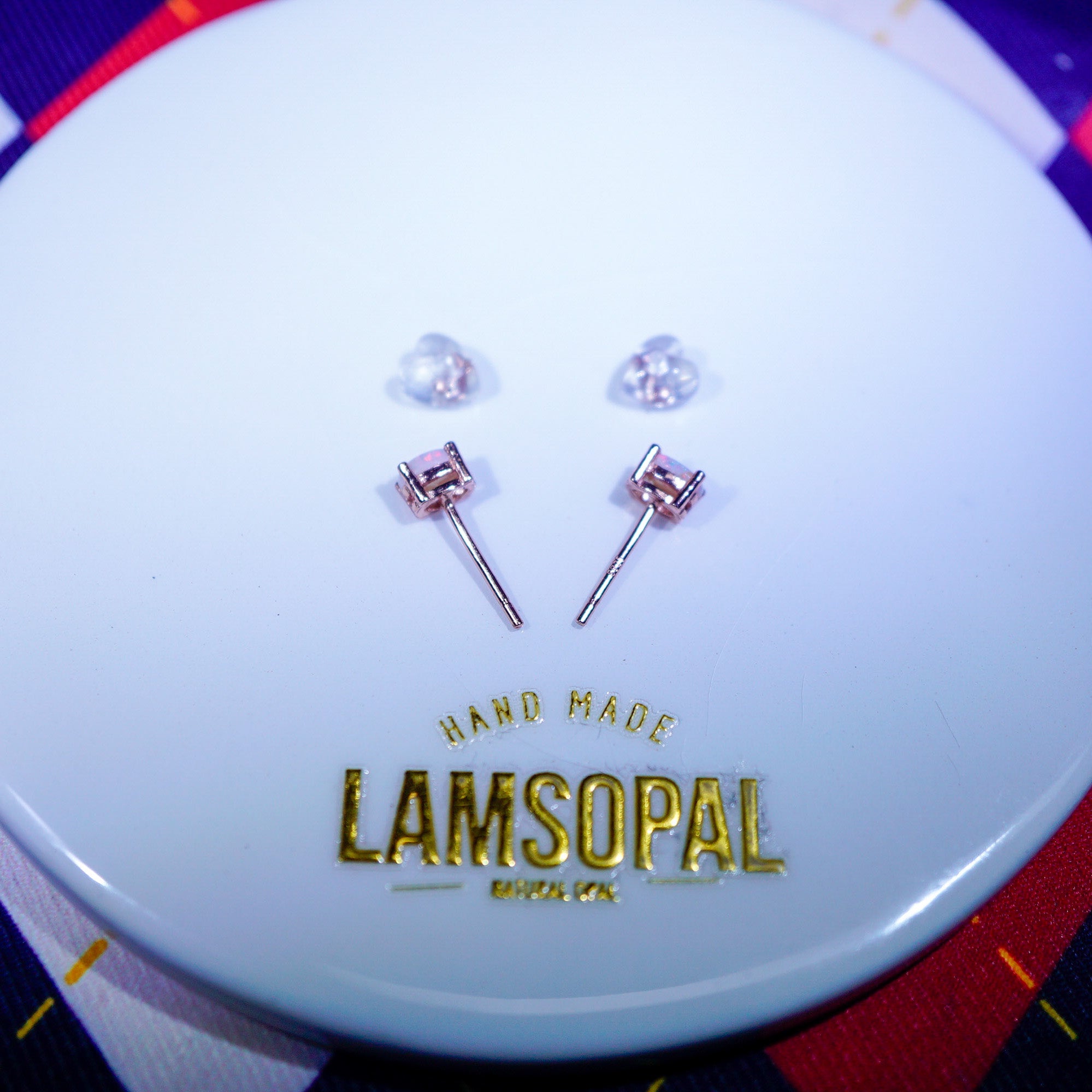 A pair of rose gold stud earrings with clear gemstones, displayed on a white circular branded stand that reads 'HAND MADE LAMSOPAL'.