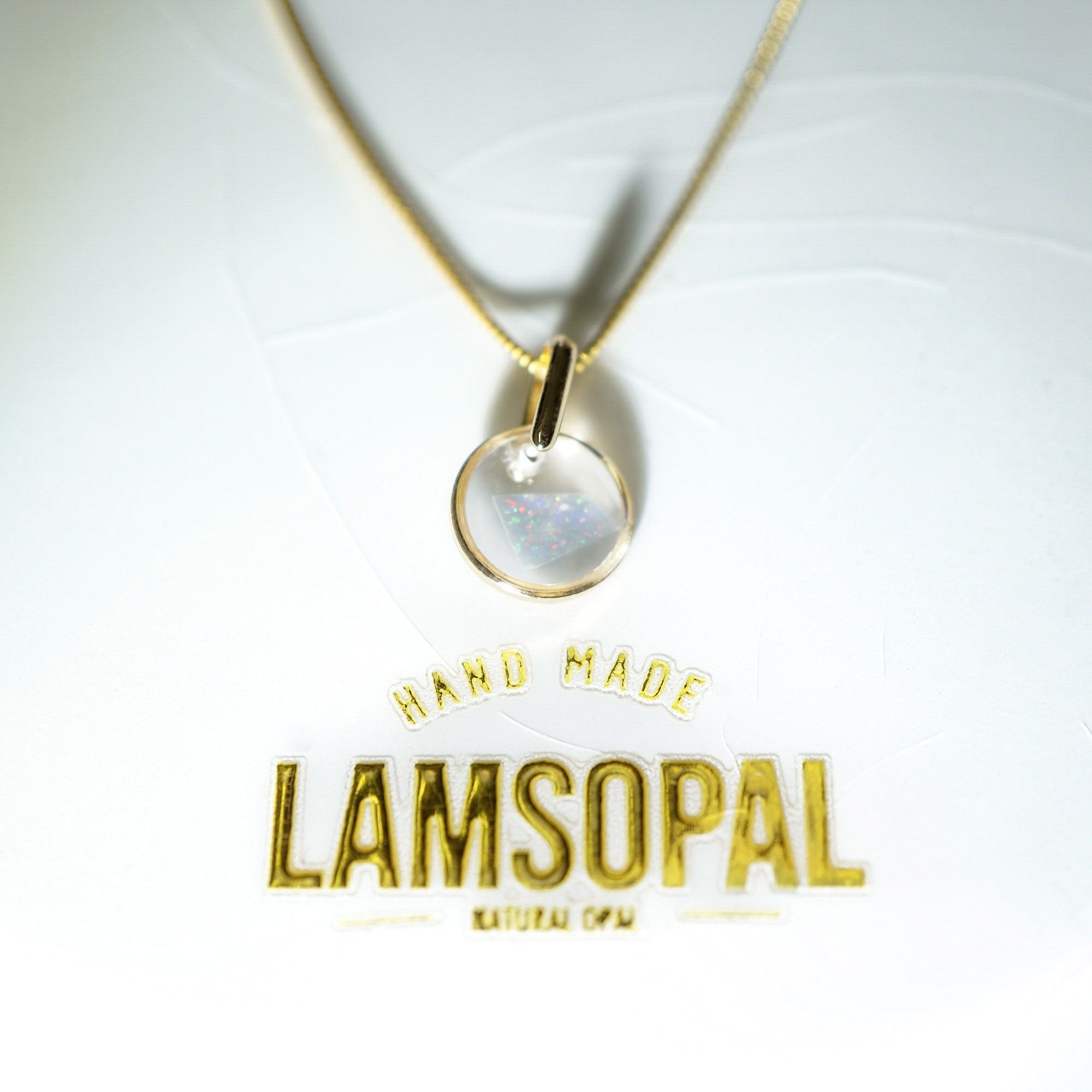 A gold necklace with a round bezel-set natural opal pendant, displayed on a white background with gold foil branding that reads 'HAND MADE LAMSOPAL'.