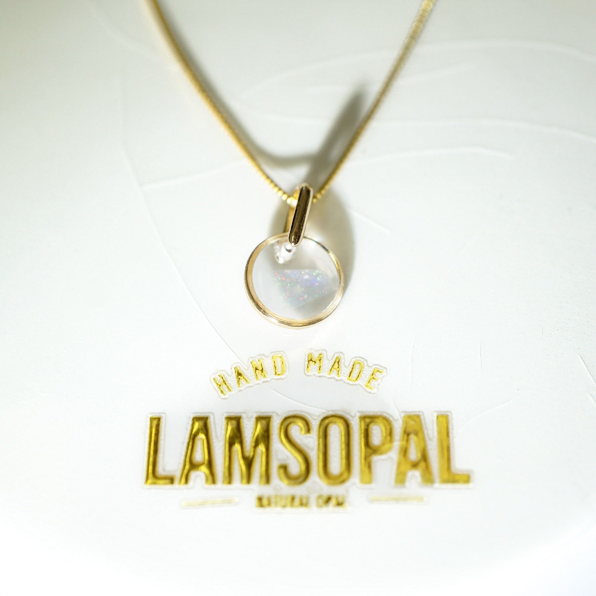 A gold chain necklace with a circular pendant containing an iridescent moonstone, displayed on a white textured background with a gold 'LAMSOPAL' brand logo below.