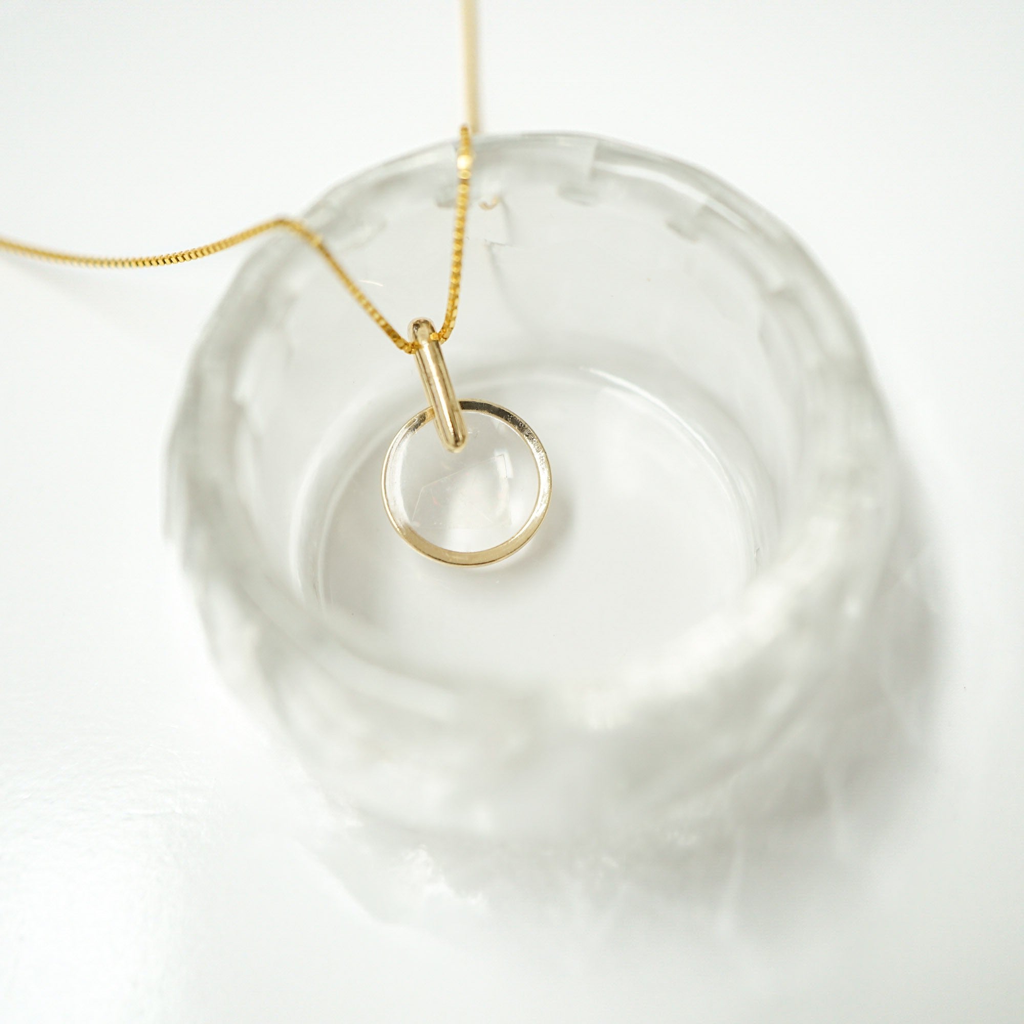 A delicate gold chain necklace with a minimalist, open circular ring pendant, displayed inside a textured clear glass bowl on a white background.