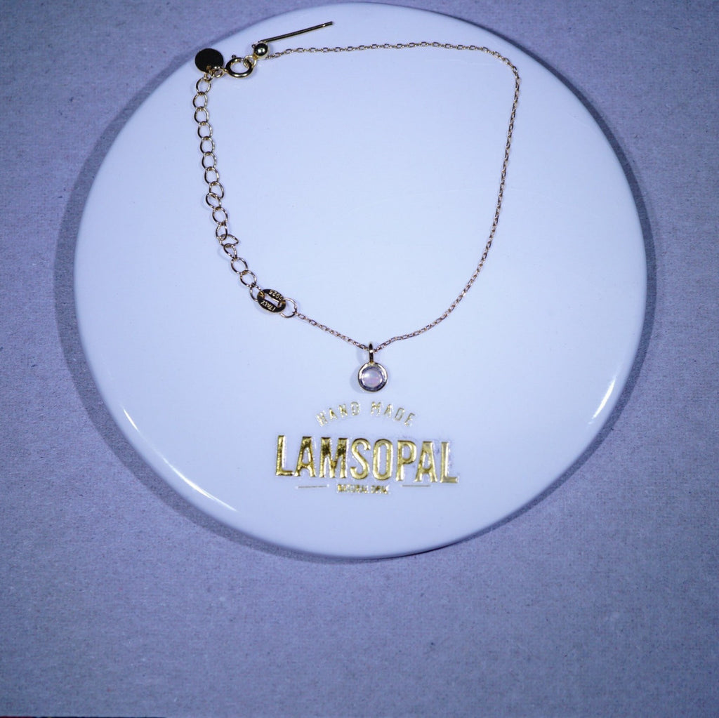 A gold-tone chain bracelet with a small oval bezel-set clear stone pendant, displayed on a white circular branded card that reads 'LAMSOPAL'.