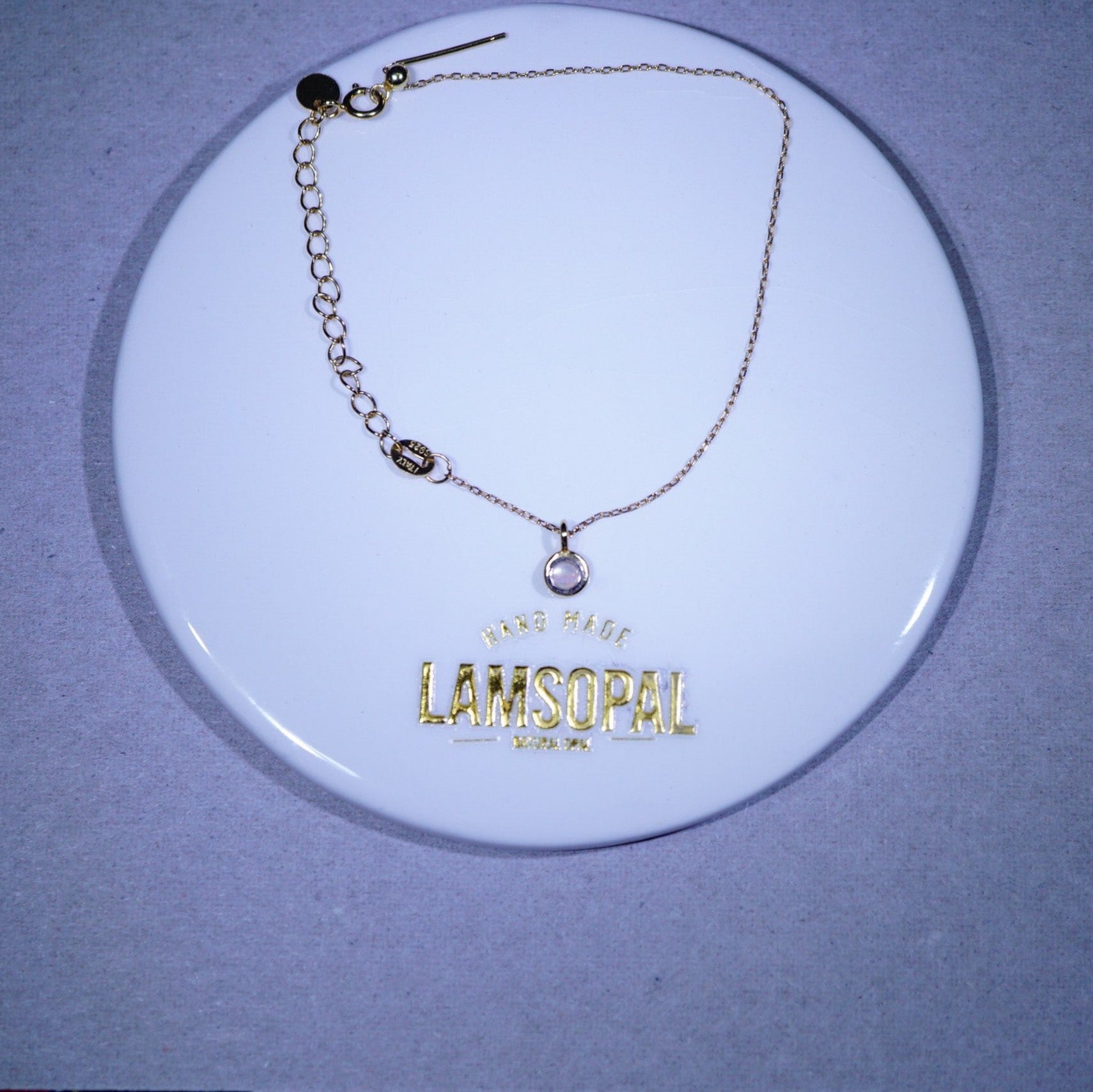 A gold-tone chain bracelet with a small oval bezel-set clear stone pendant, displayed on a white circular branded card that reads 'LAMSOPAL'.
