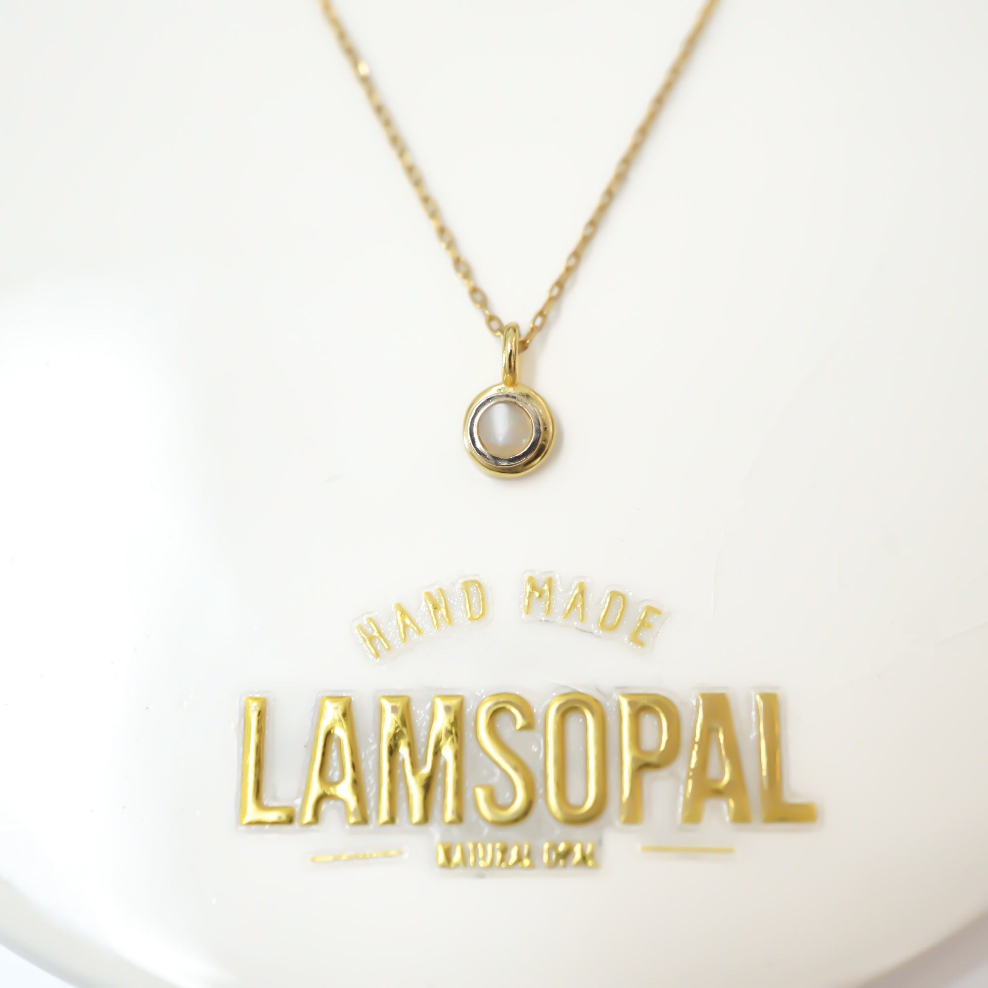 A gold chain necklace with a small circular pendant featuring a white inlay, displayed on a white surface with a gold 'LAMSOPAL' brand plaque below.
