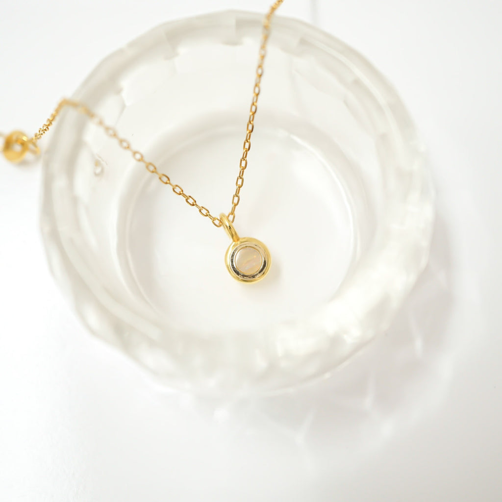 A delicate gold chain necklace with a small circular pendant featuring a pale pink stone, displayed on a textured clear glass dish against a bright white background.