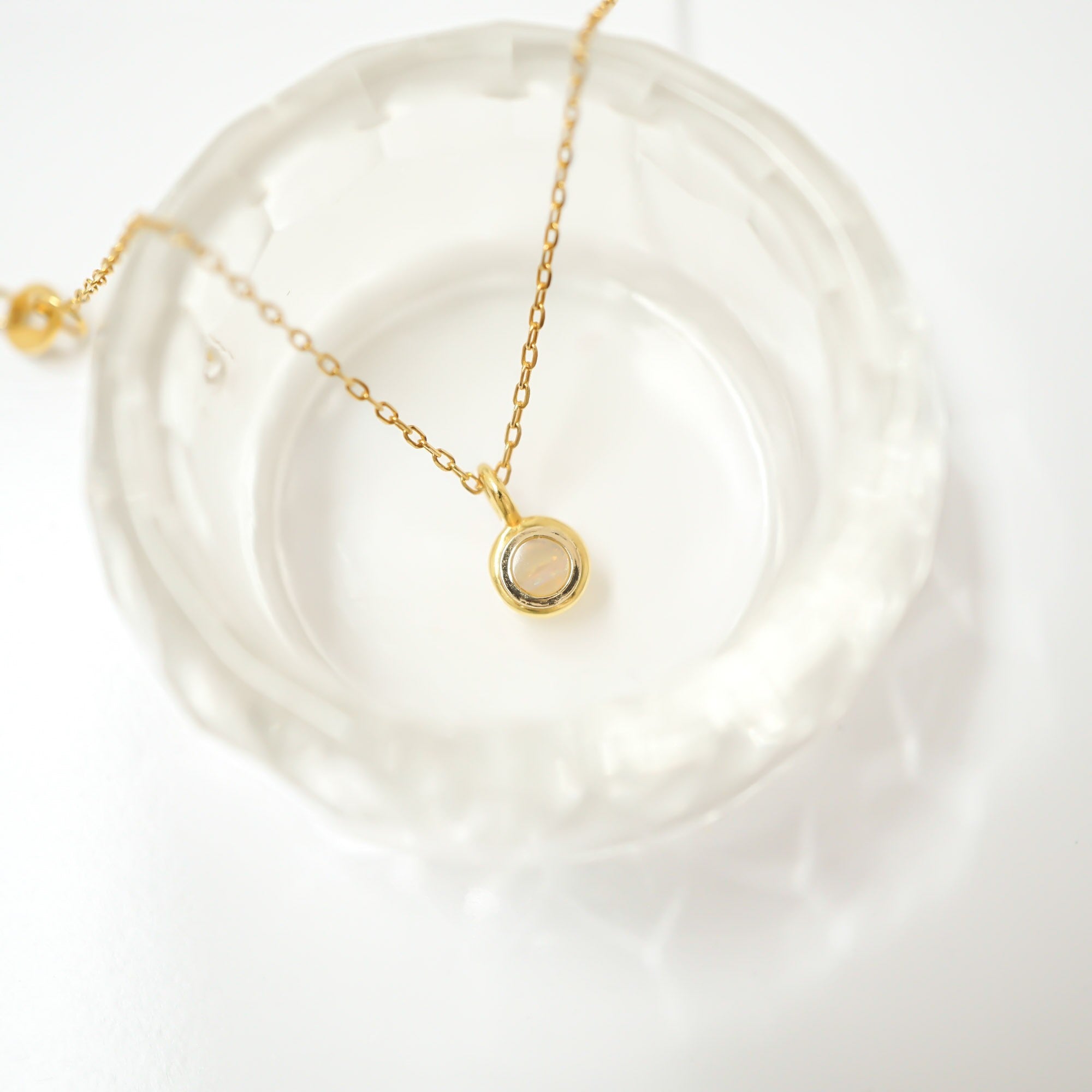 A delicate gold chain necklace with a small circular pendant featuring a pale pink stone, displayed on a textured clear glass dish against a bright white background.