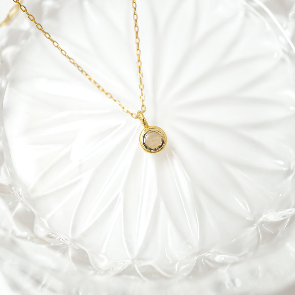 A delicate gold chain necklace with a small circular pendant resting on a white textured glass plate.