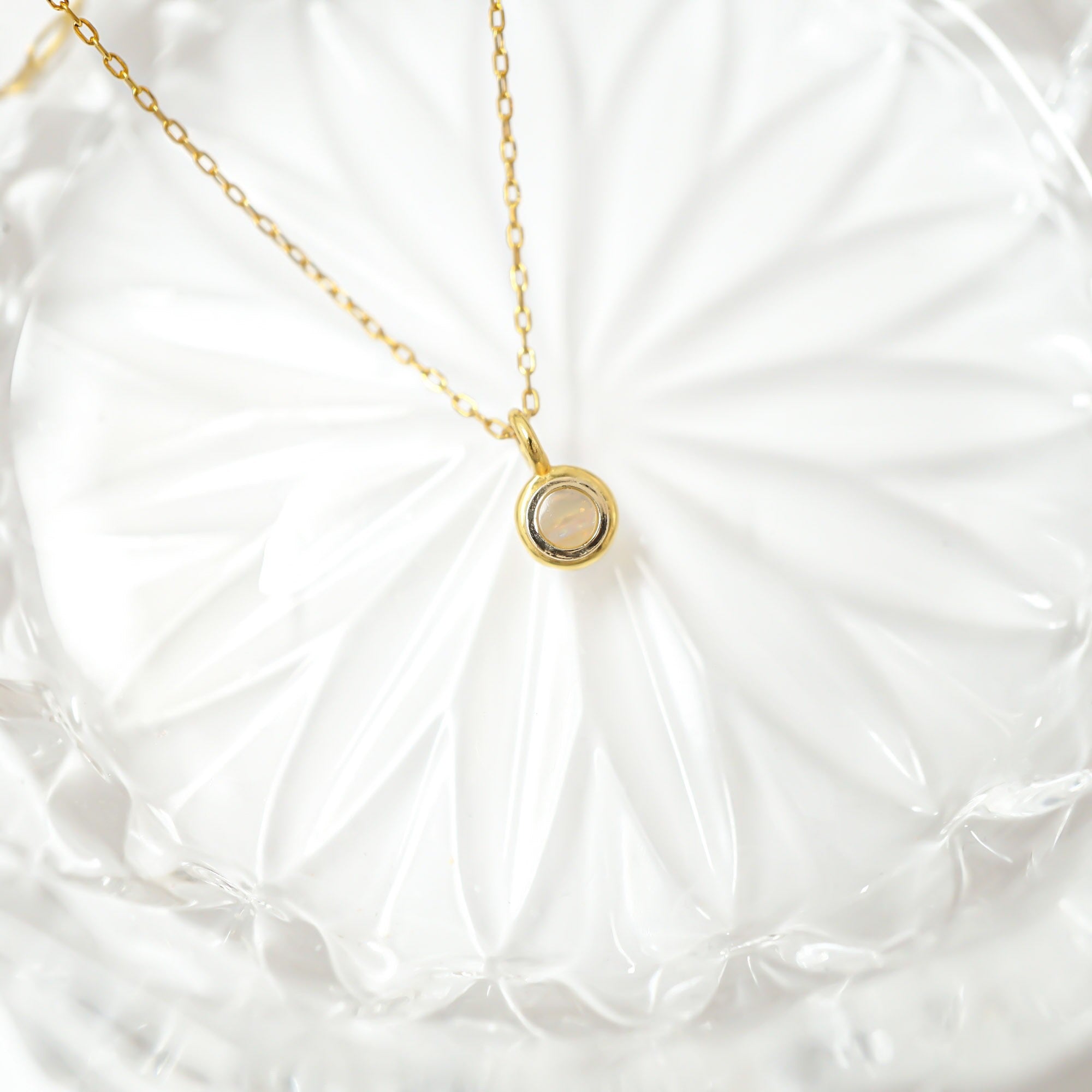 A delicate gold chain necklace with a small circular pendant resting on a white textured glass plate.