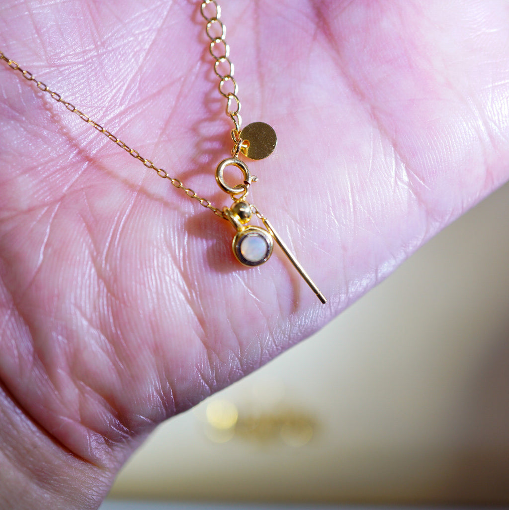 A delicate gold chain necklace with a small round gold disc charm, a ring connector, and a pendant featuring a white circular stone and a thin gold bar, resting on a person's palm.