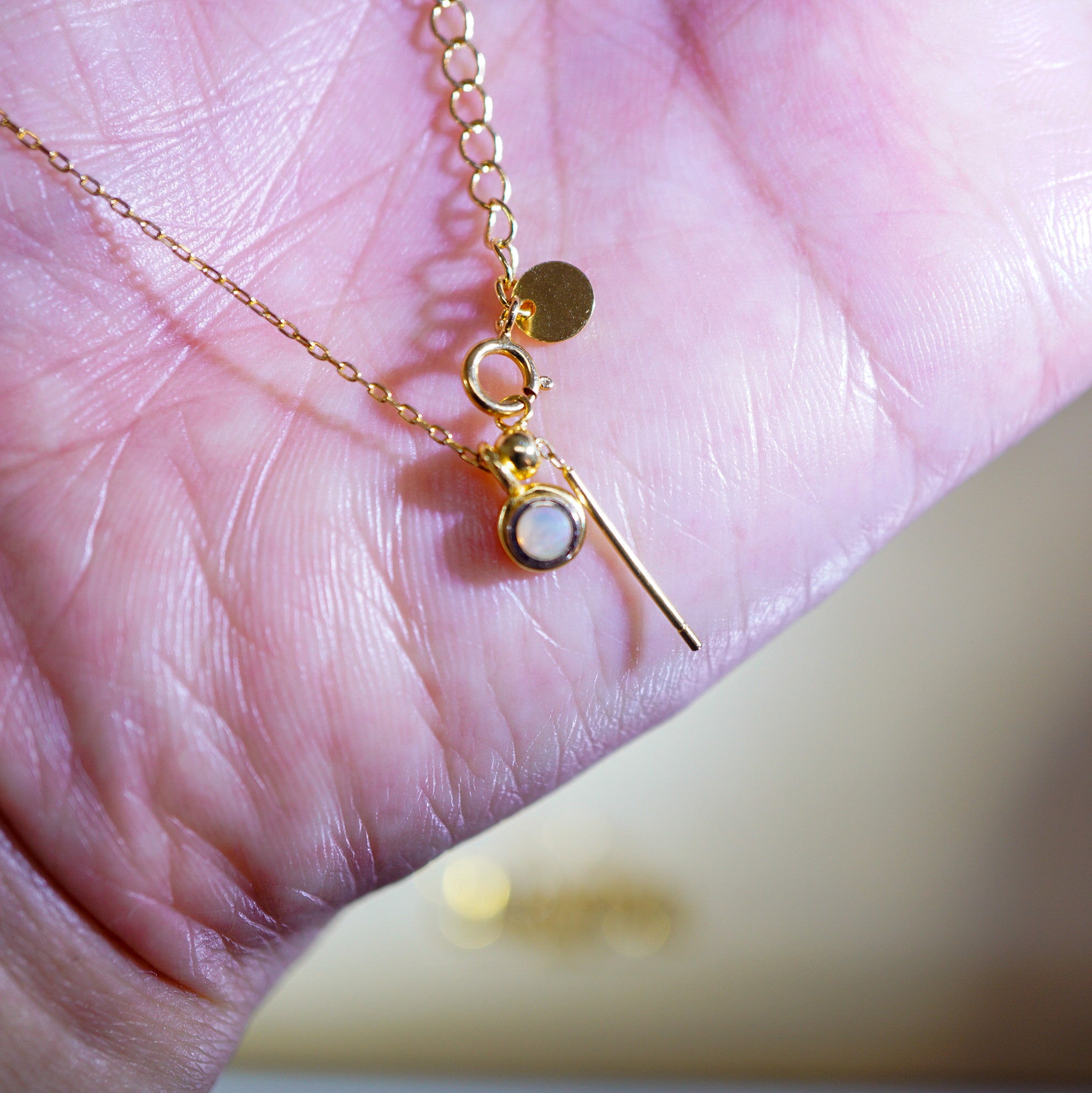 A delicate gold chain necklace with a small round gold disc charm, a ring connector, and a pendant featuring a white circular stone and a thin gold bar, resting on a person's palm.