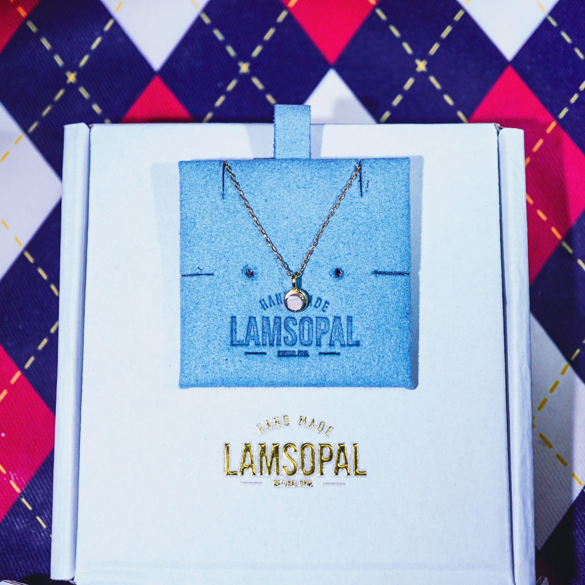 A delicate gold chain necklace with a small round pendant displayed on a blue card inside an open white Lamsopal gift box, set against a red, navy, and white argyle patterned fabric background.