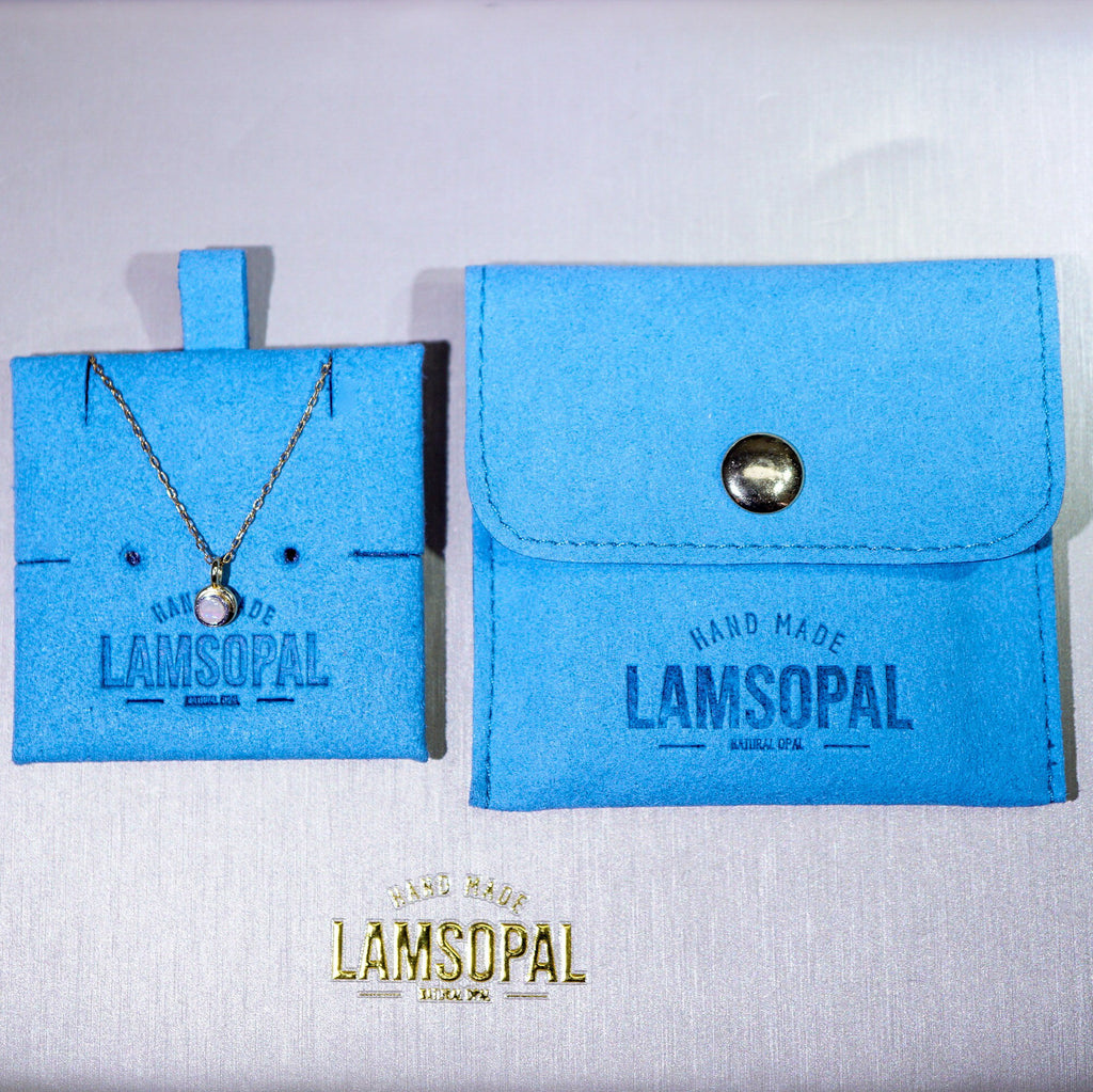 A Lamsopal necklace displayed on a bright blue jewelry card next to a matching blue pouch with a snap closure, on a light fabric background. The Lamsopal brand name is embossed on the card and printed on the pouch, with a gold Lamsopal logo at the bottom.