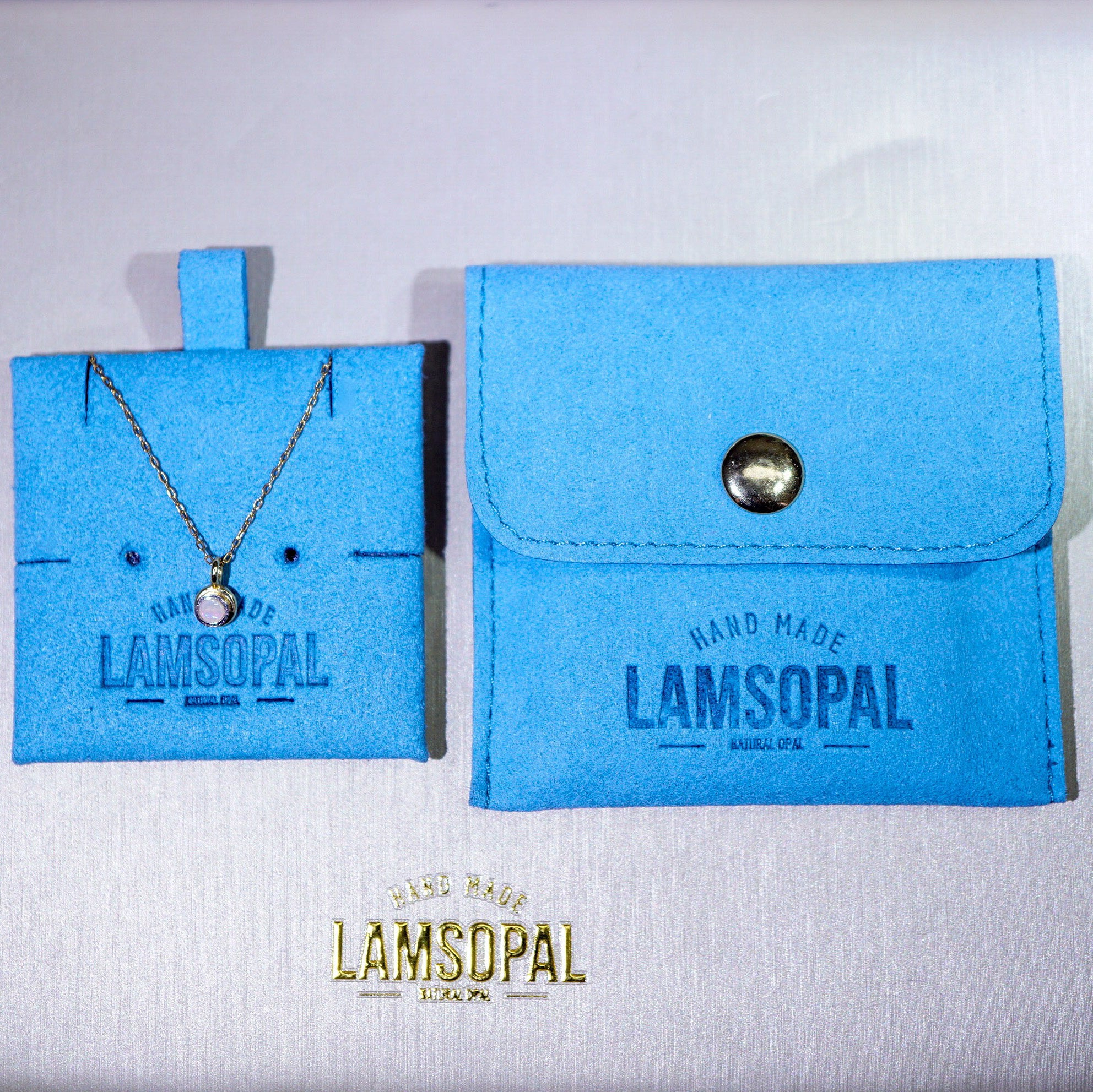 A Lamsopal necklace displayed on a bright blue jewelry card next to a matching blue pouch with a snap closure, on a light fabric background. The Lamsopal brand name is embossed on the card and printed on the pouch, with a gold Lamsopal logo at the bottom.
