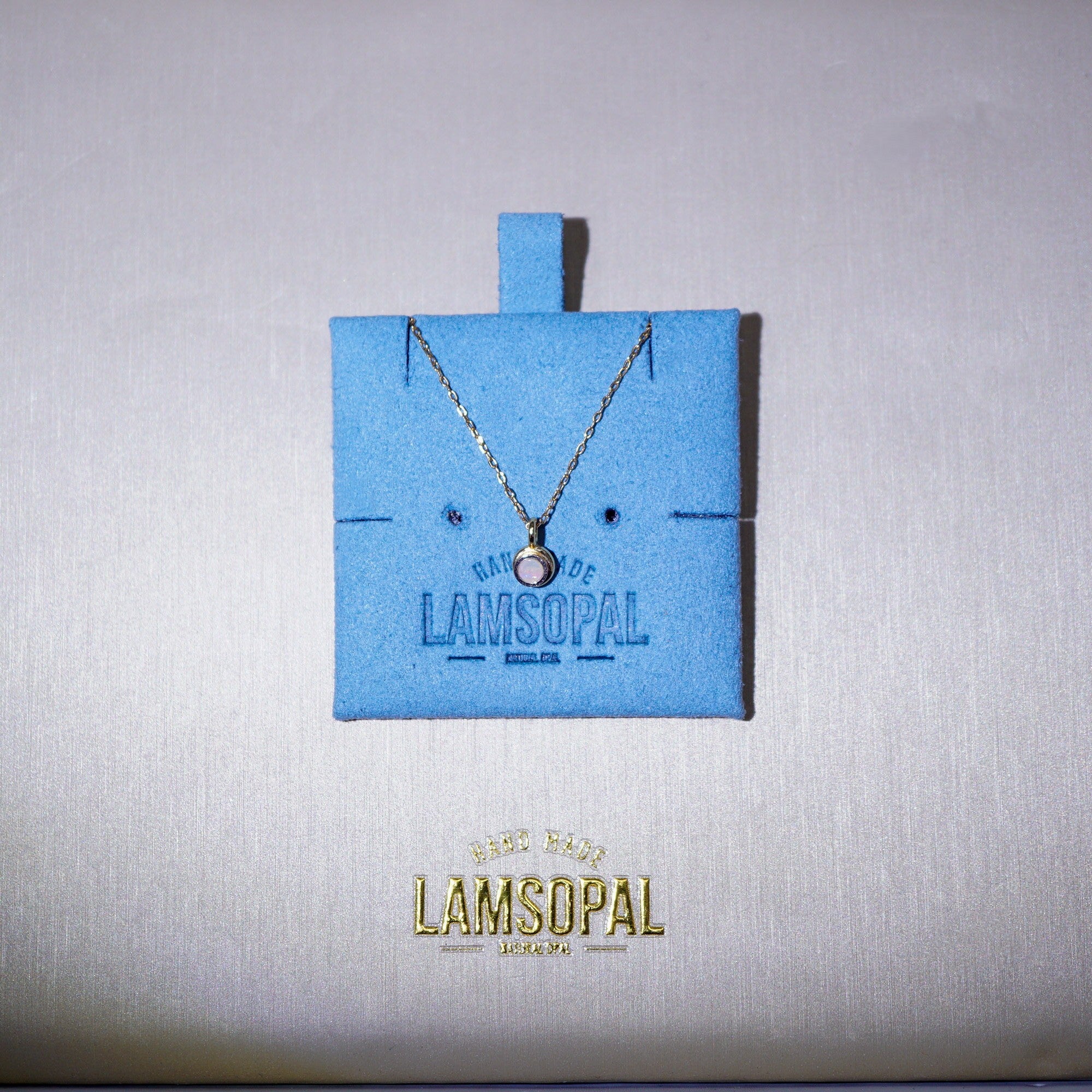 A delicate silver chain necklace with a small round pendant displayed on a bright blue Lamsopal jewelry card, set against a textured light gray background with the gold Lamsopal logo below.