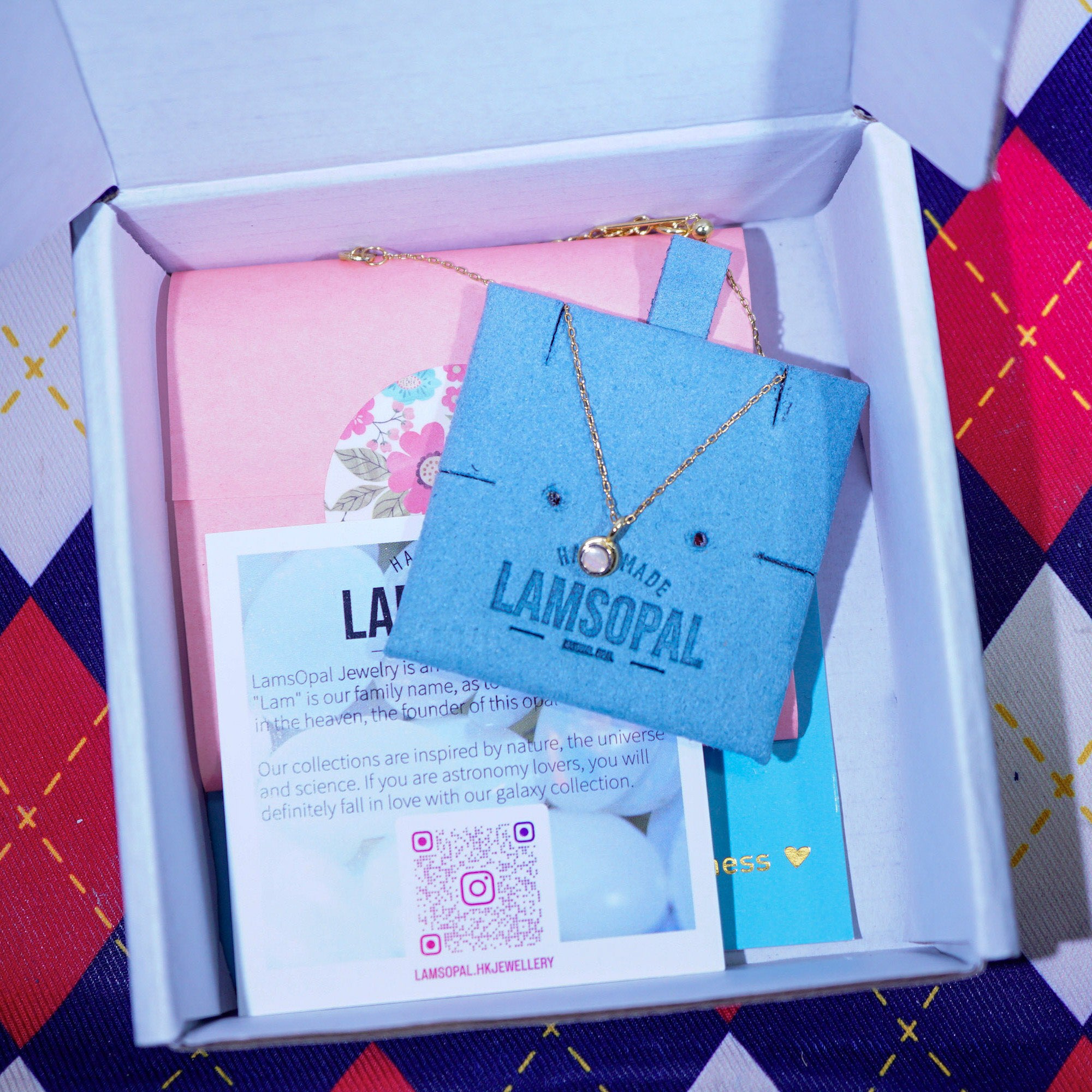 A gold necklace with a small round pendant displayed on a blue Lamsopal card inside an open gift box, with a pink pouch and printed cards visible around it.