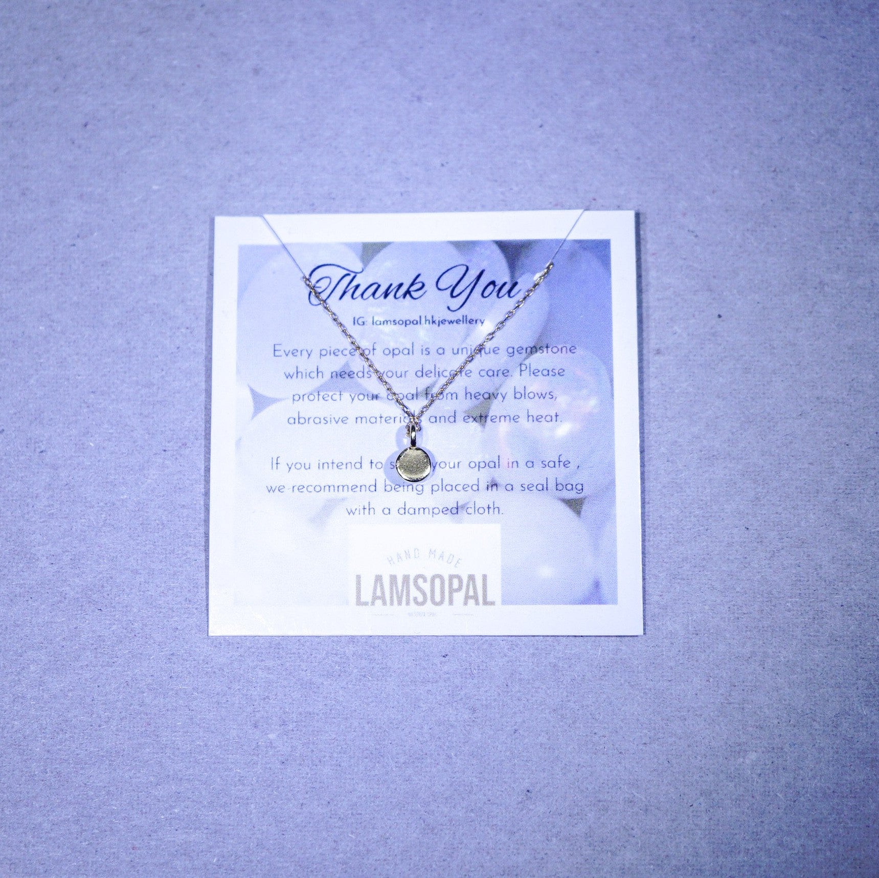 A delicate silver-toned necklace with a small oval gemstone pendant is displayed on a printed card that reads 'Thank You' and 'LAMSOPAL', set against a textured light purple background.