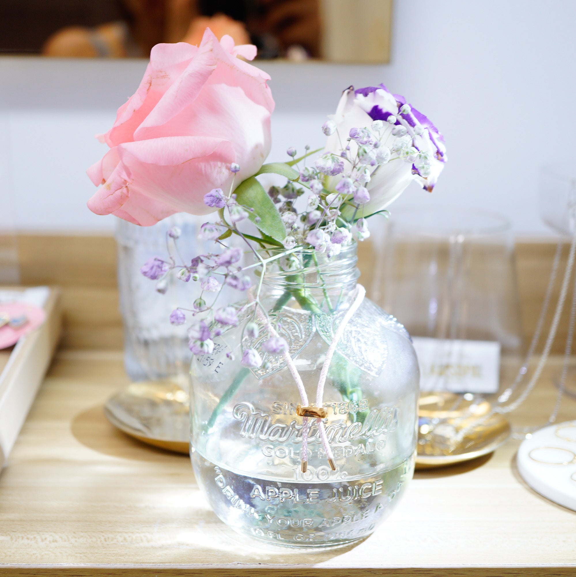 A clear glass jar vase with embossed text, filled with water and decorated with a pink rose, a purple and white flower, and small lavender sprigs, displayed on a light wooden surface.