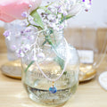 A delicate light pink beaded necklace with a blue faceted bead pendant, draped over a vintage embossed glass mason jar vase filled with pink and white flowers.