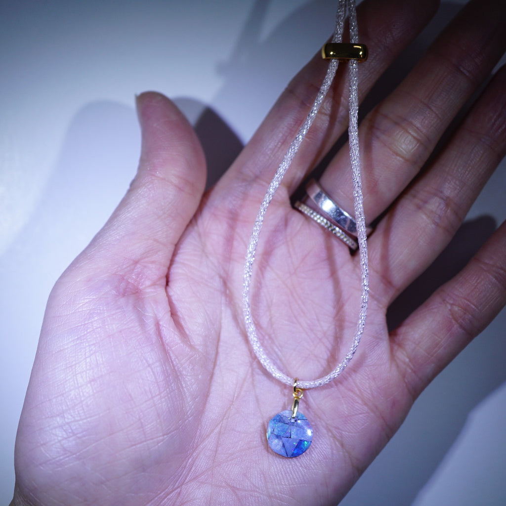 A delicate necklace with a textured white cord and a round blue pendant set in gold, held in a person's open hand.