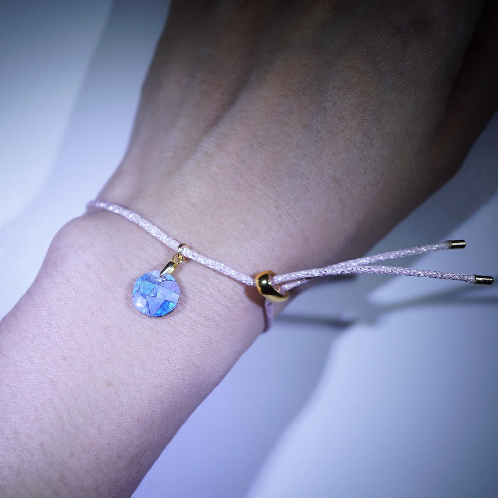 A close-up of a delicate bracelet worn on a wrist, featuring a light pink glitter cord, a small gold clasp, and a faceted light blue crystal charm.