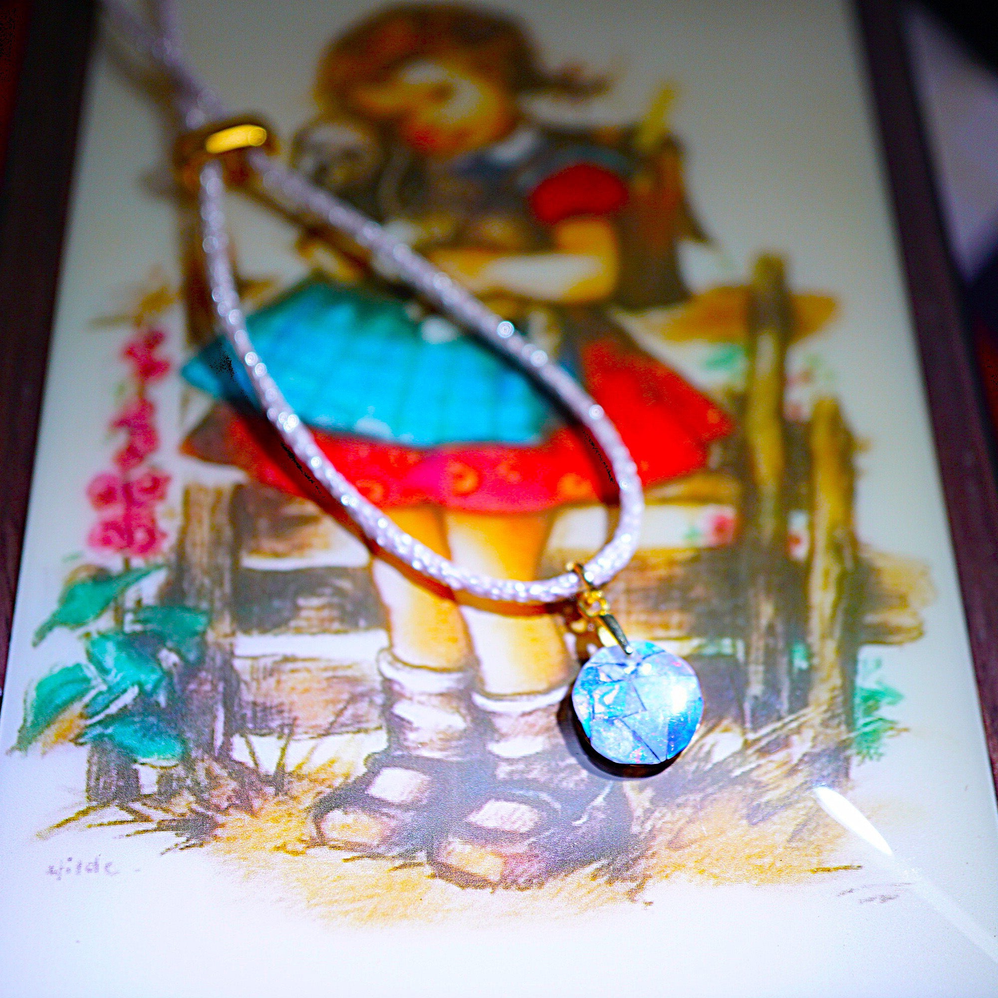 A close-up of a beaded necklace with a round blue pendant resting on a vintage-style illustrated card featuring a girl in a red and blue dress.