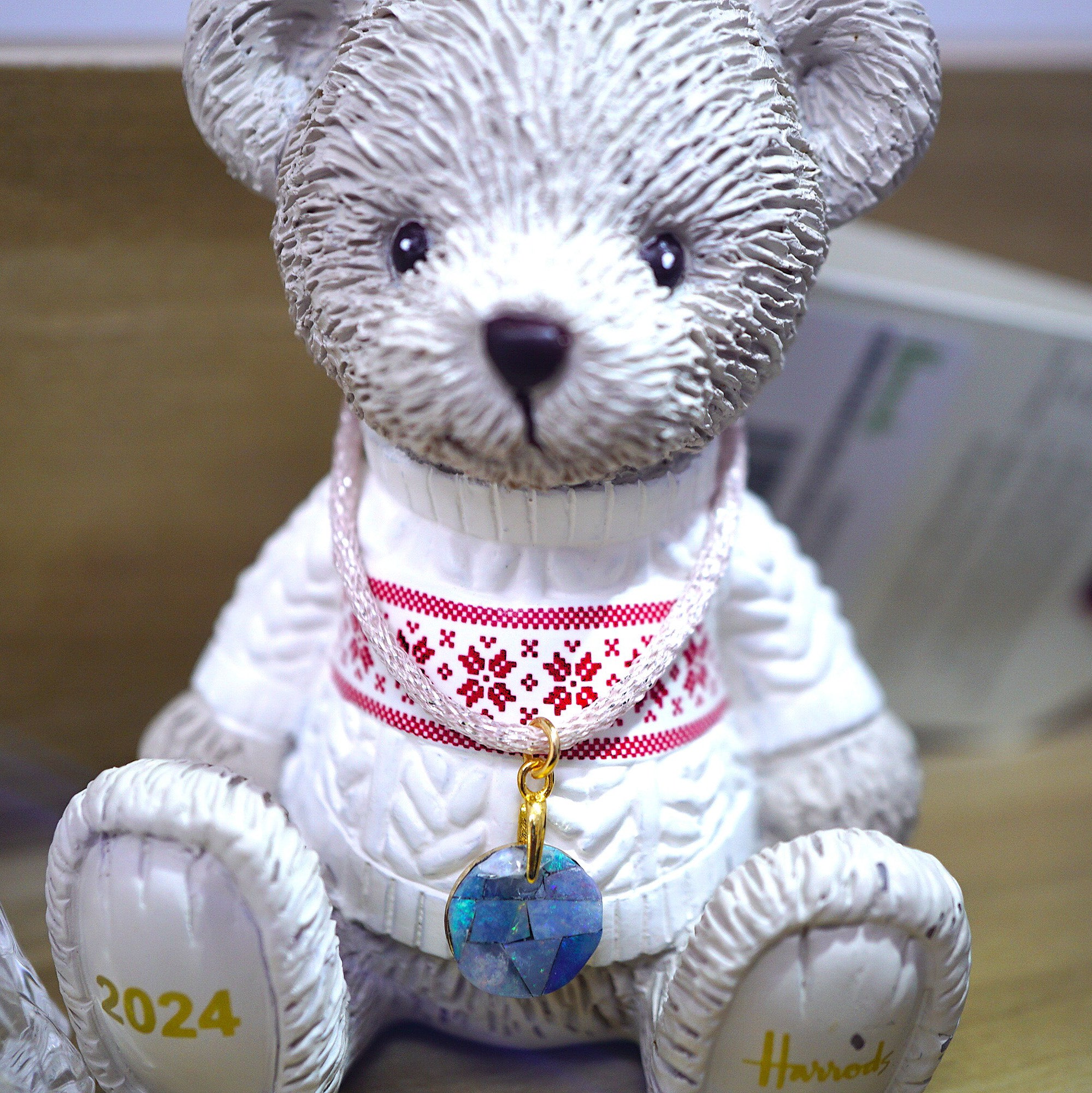 A white teddy bear figurine wearing a white knit sweater with a red Nordic pattern and a blue opal circular pendant. The bear has '2024' printed on one paw and the name 'Harriet' printed on the other, set against a blurred indoor background.