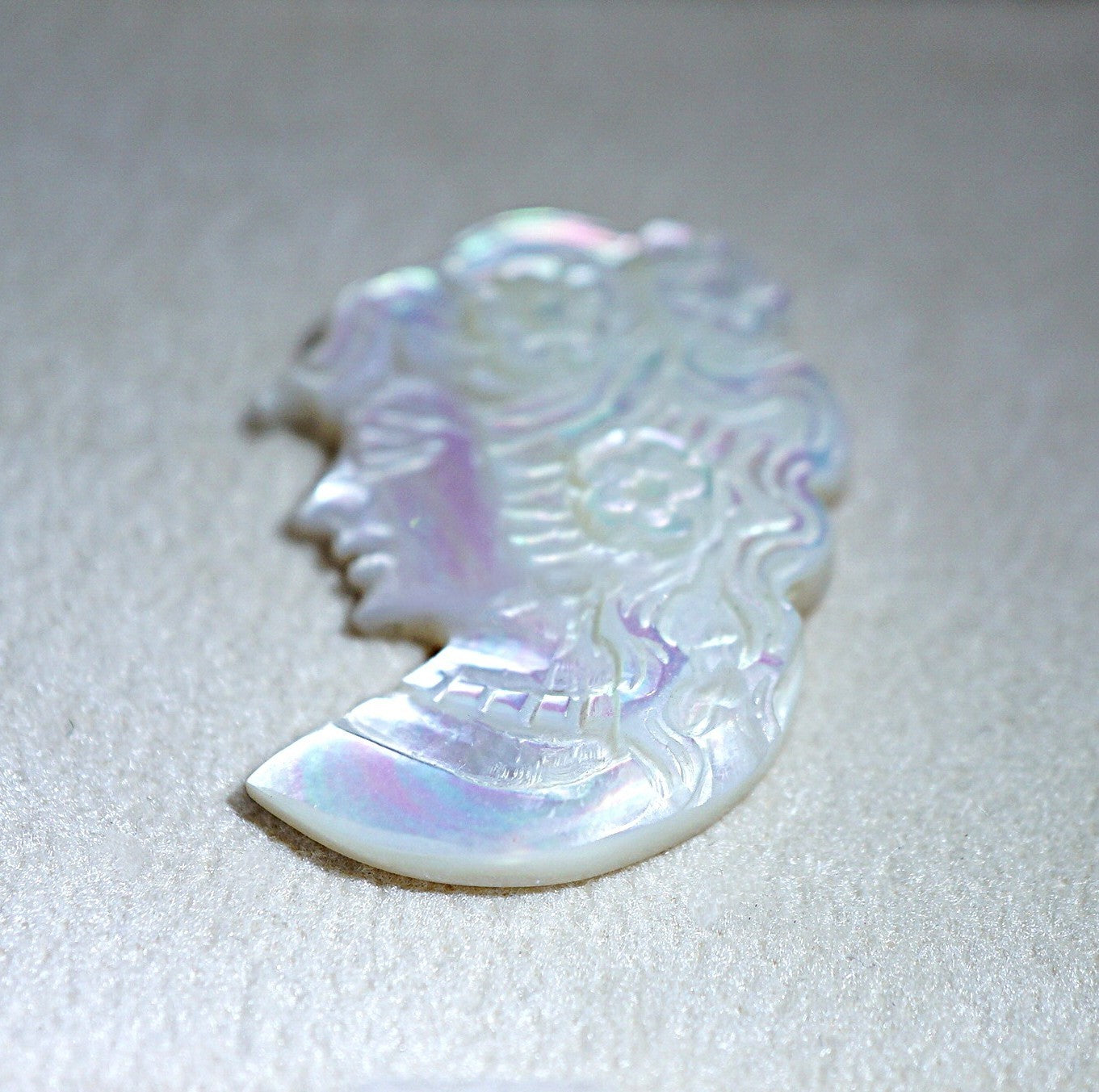 A carved iridescent shell pendant or charm with a profile face design, displayed on a textured light gray background.