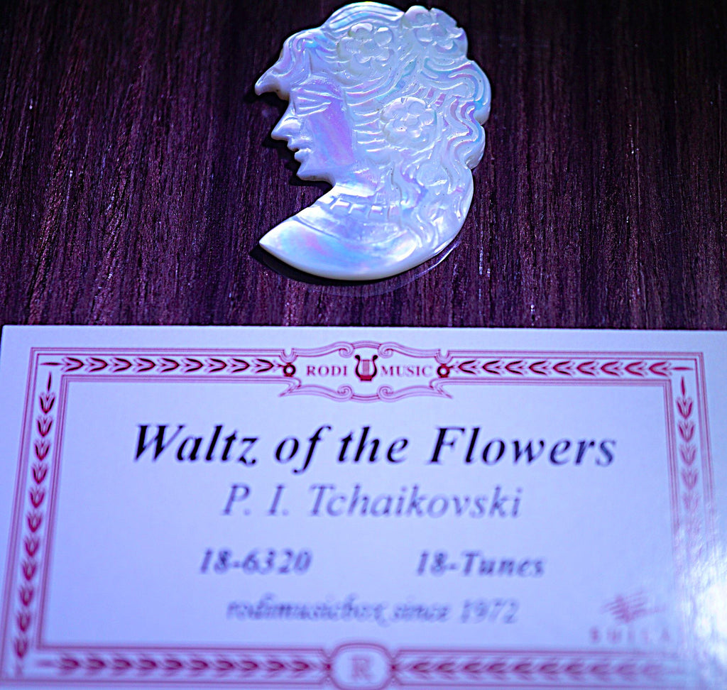 An iridescent white carved shell or pearl-like ornament shaped like a profile portrait, placed above a printed music card titled 'Waltz of the Flowers' by P. I.