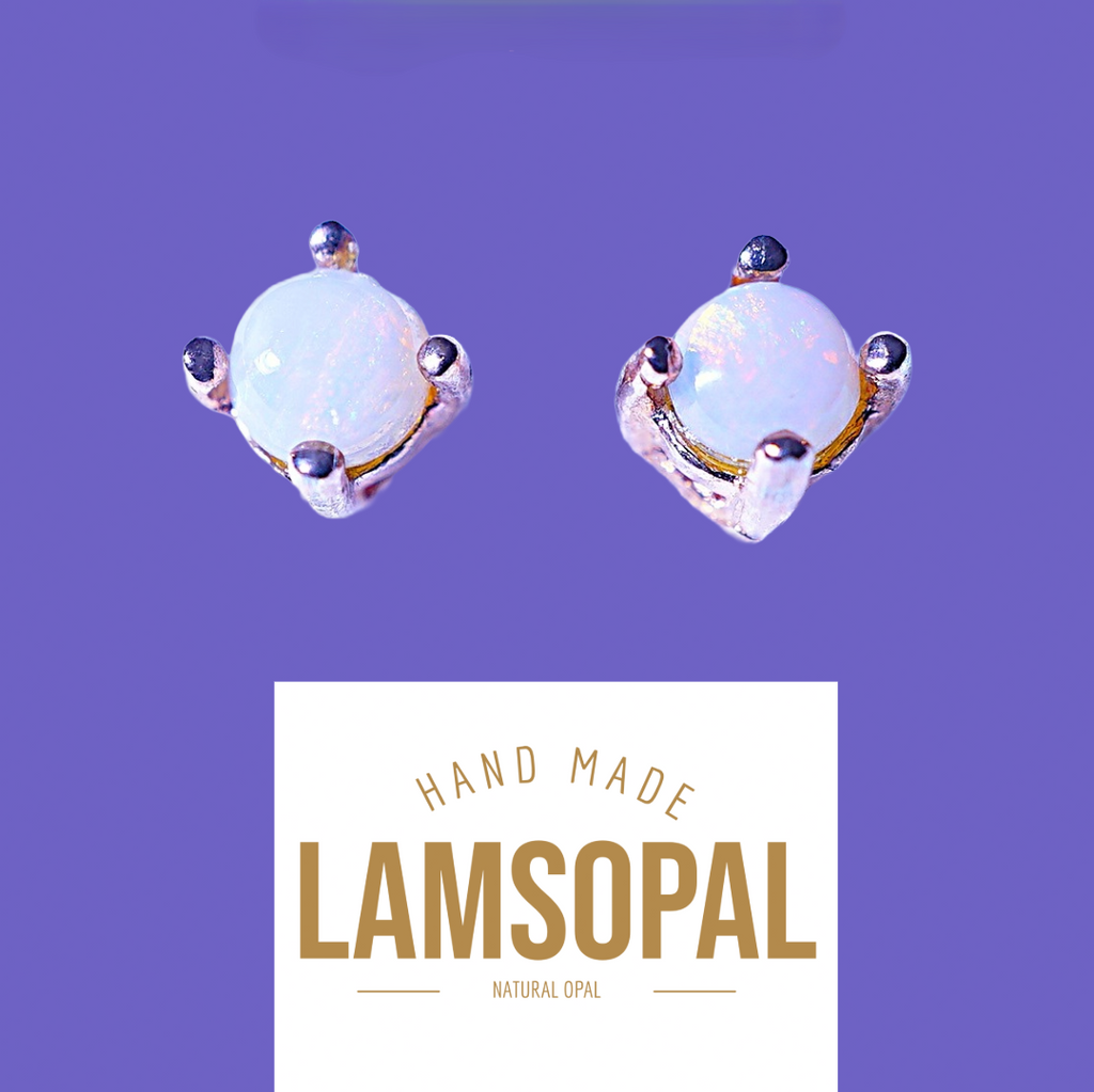 A pair of handmade Lamsopal natural opal stud earrings with silver settings and small dark accent stones, shown above a white label that reads 'HAND MADE LAMSOPAL NATURAL OPAL' on a solid purple background.
