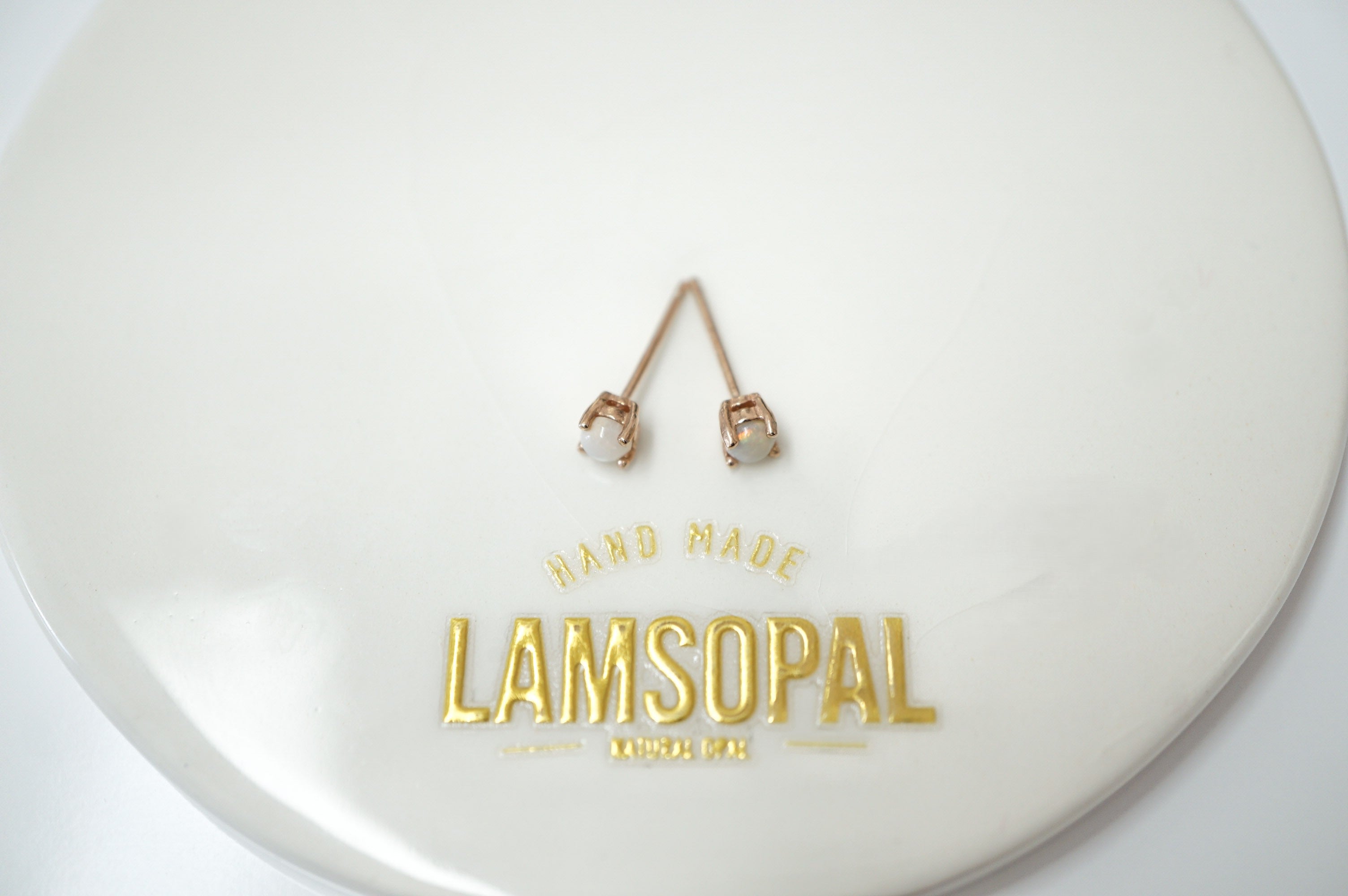 A pair of rose gold stud earrings with clear gemstones, displayed on a white circular stand with gold foil branding that reads 'HAND MADE LAMSOPAL'.