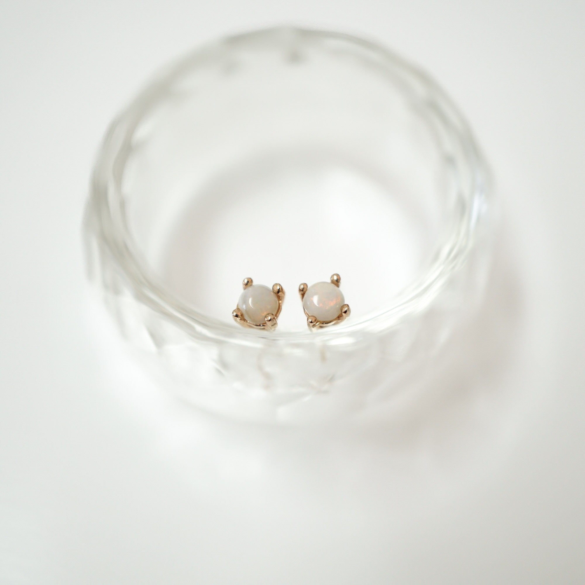 A pair of small gold stud earrings with oval milky white stones, displayed inside a clear textured glass dish on a white background.