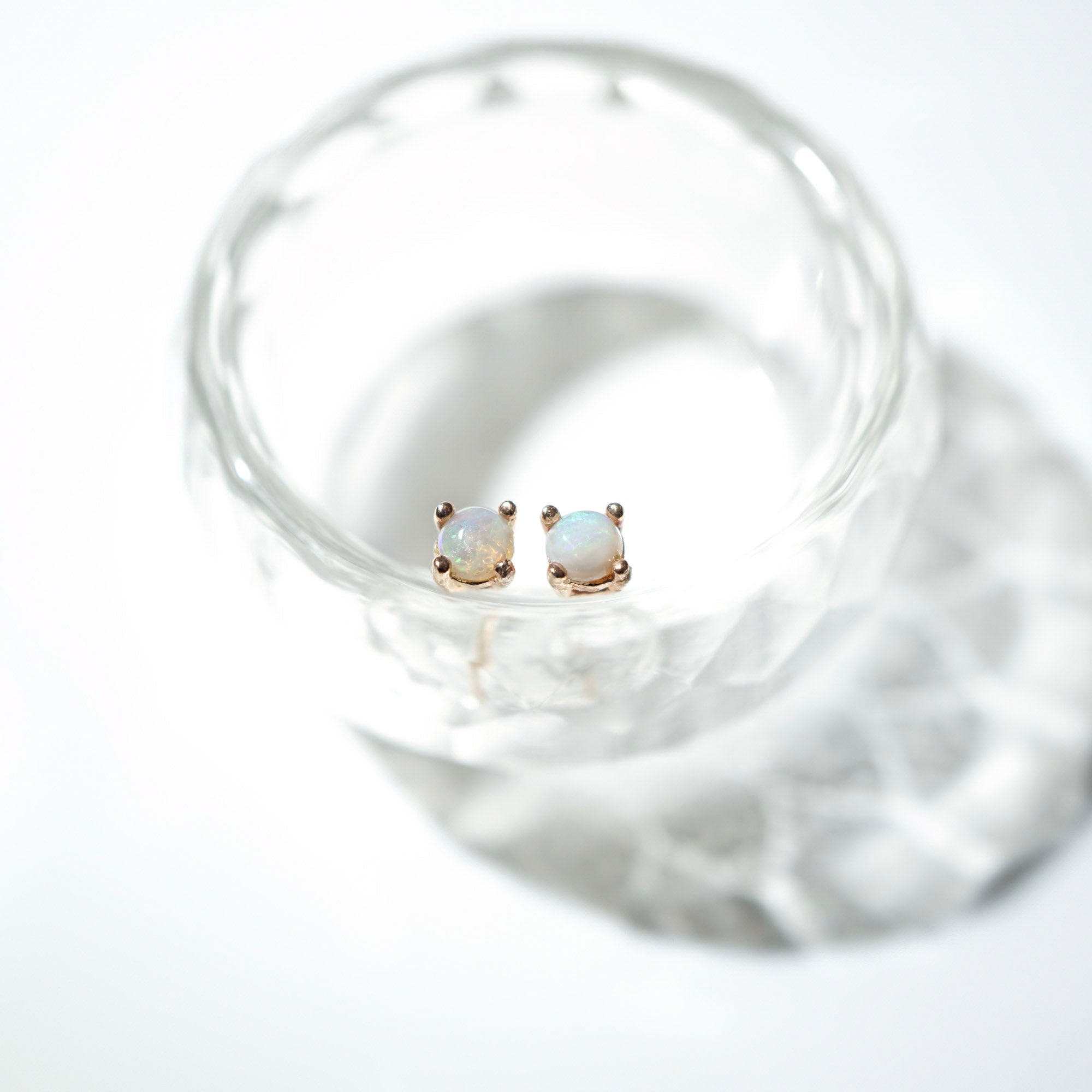 A pair of small square stud earrings with iridescent white stones in gold settings, resting inside a clear textured glass on a white background.