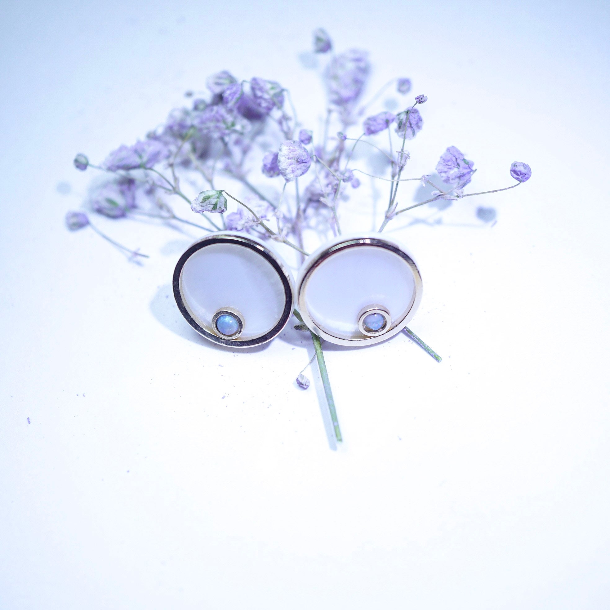 A pair of round stud earrings with silver-toned metal rims and pale blue oval centers, styled with dried lavender flowers on a white background.