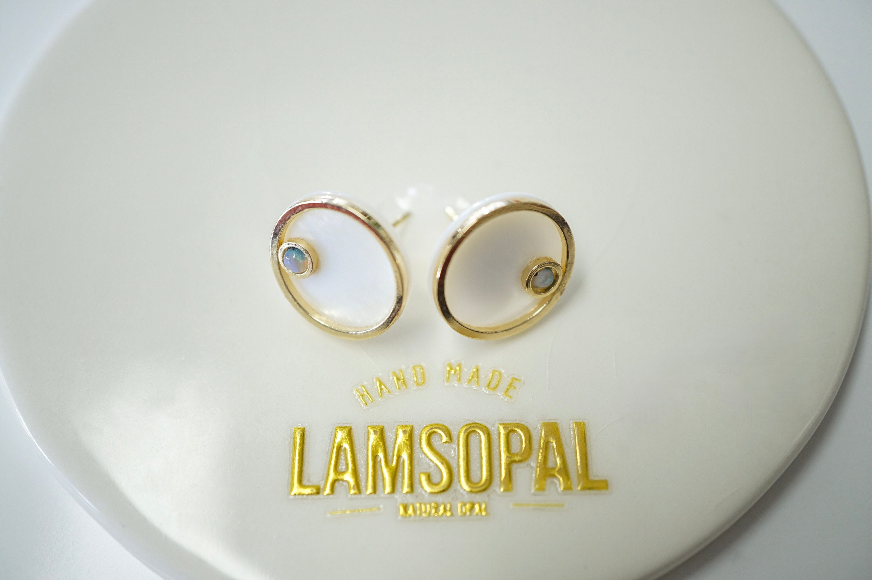 A pair of gold-toned oval hoop earrings with small light blue gemstones, displayed on a white circular stand with the brand name LAMSOPAL printed in gold.