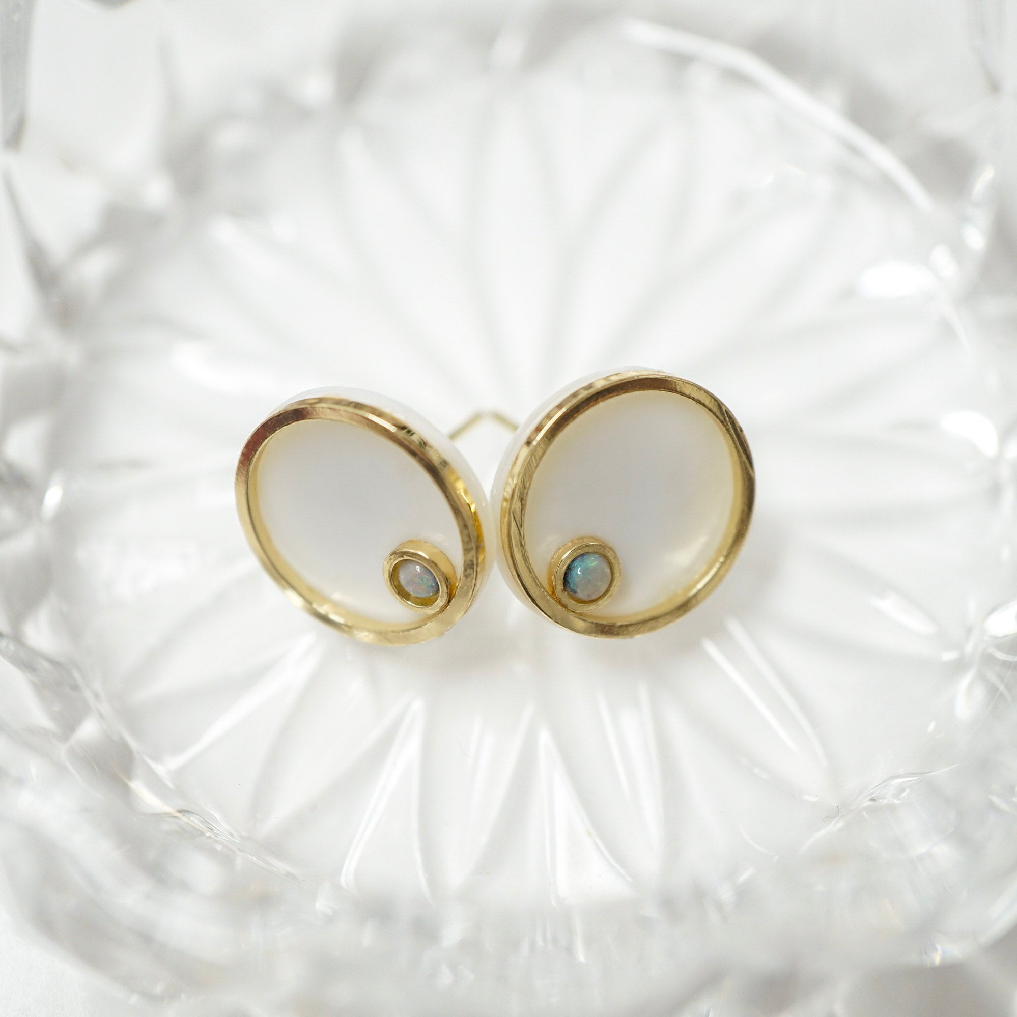 A pair of gold stud earrings with oval milky white mother-of-pearl inlays and a small iridescent blue-green stone at the bottom, displayed on a clear glass dish.